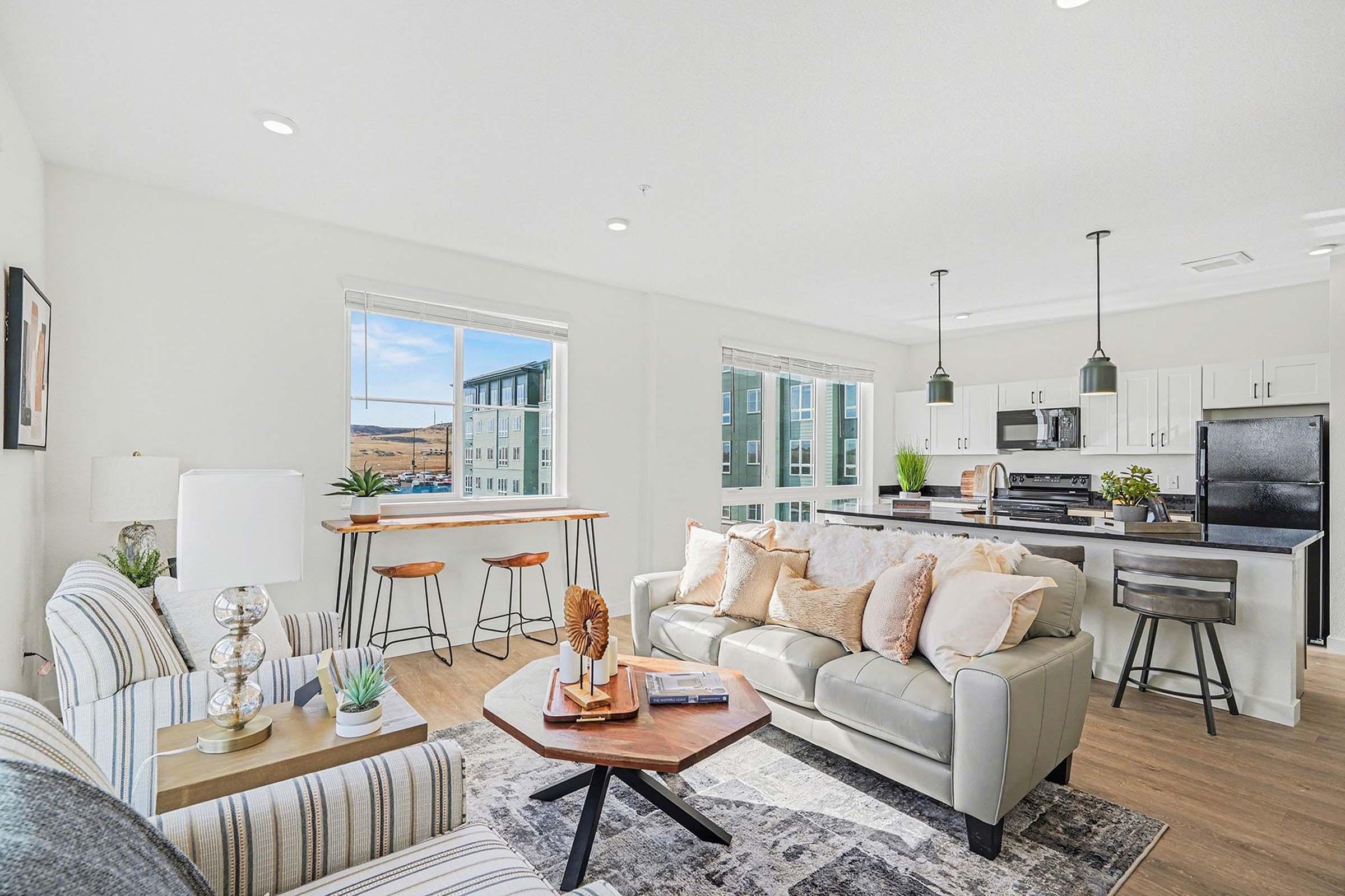 Modern open-concept living space featuring a cozy living area with a light-colored sofa and decorative pillows, a wooden coffee table, and a stylish bar with stools. The kitchen boasts sleek black appliances and white cabinetry. Large windows provide natural light and a view of the outdoors.