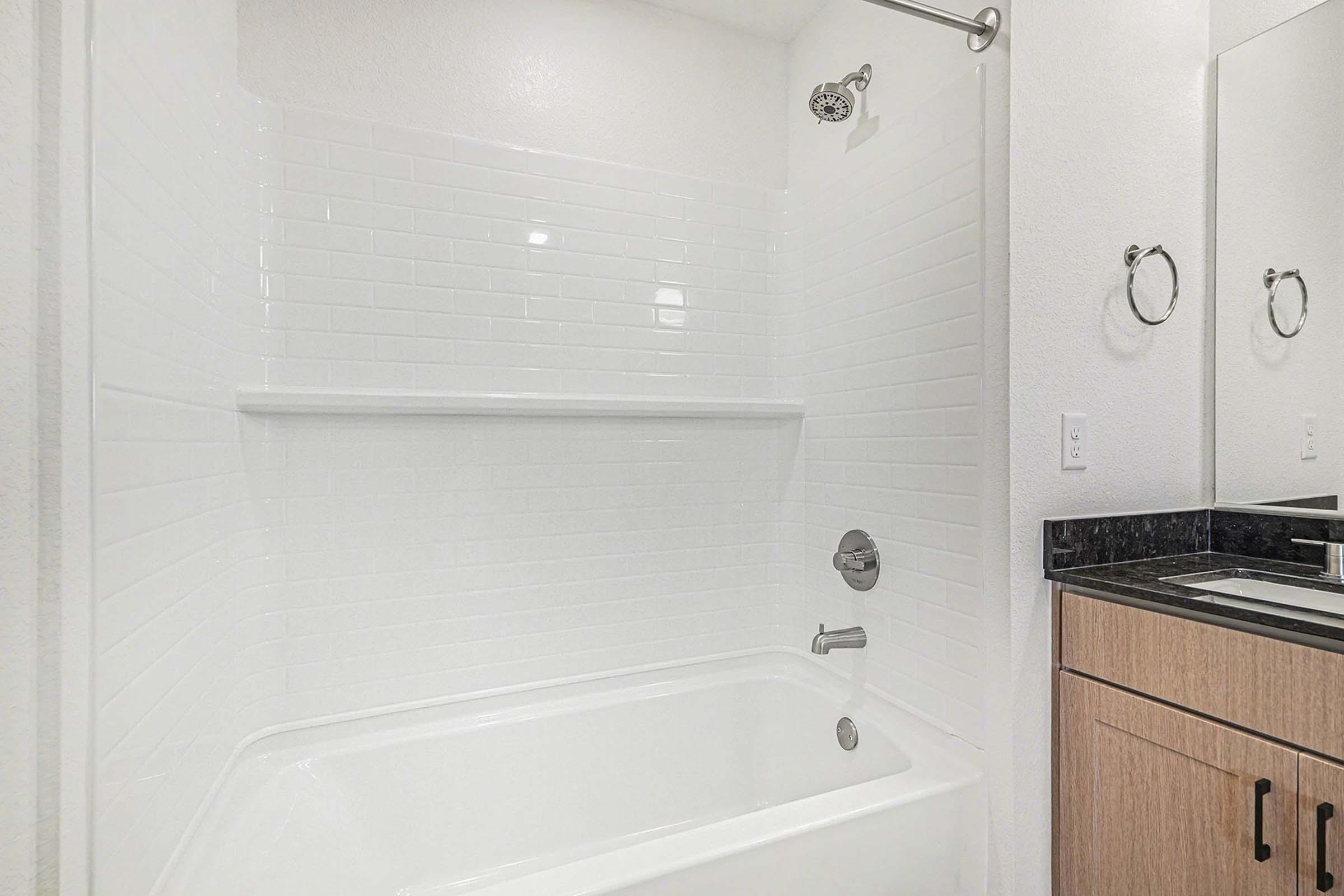 A clean and modern bathroom featuring a white bathtub and shower with a tiled wall, a chrome showerhead, and a sleek vanity with a dark countertop and wooden cabinetry. The space is well-lit with bright white walls, creating a fresh and inviting atmosphere.