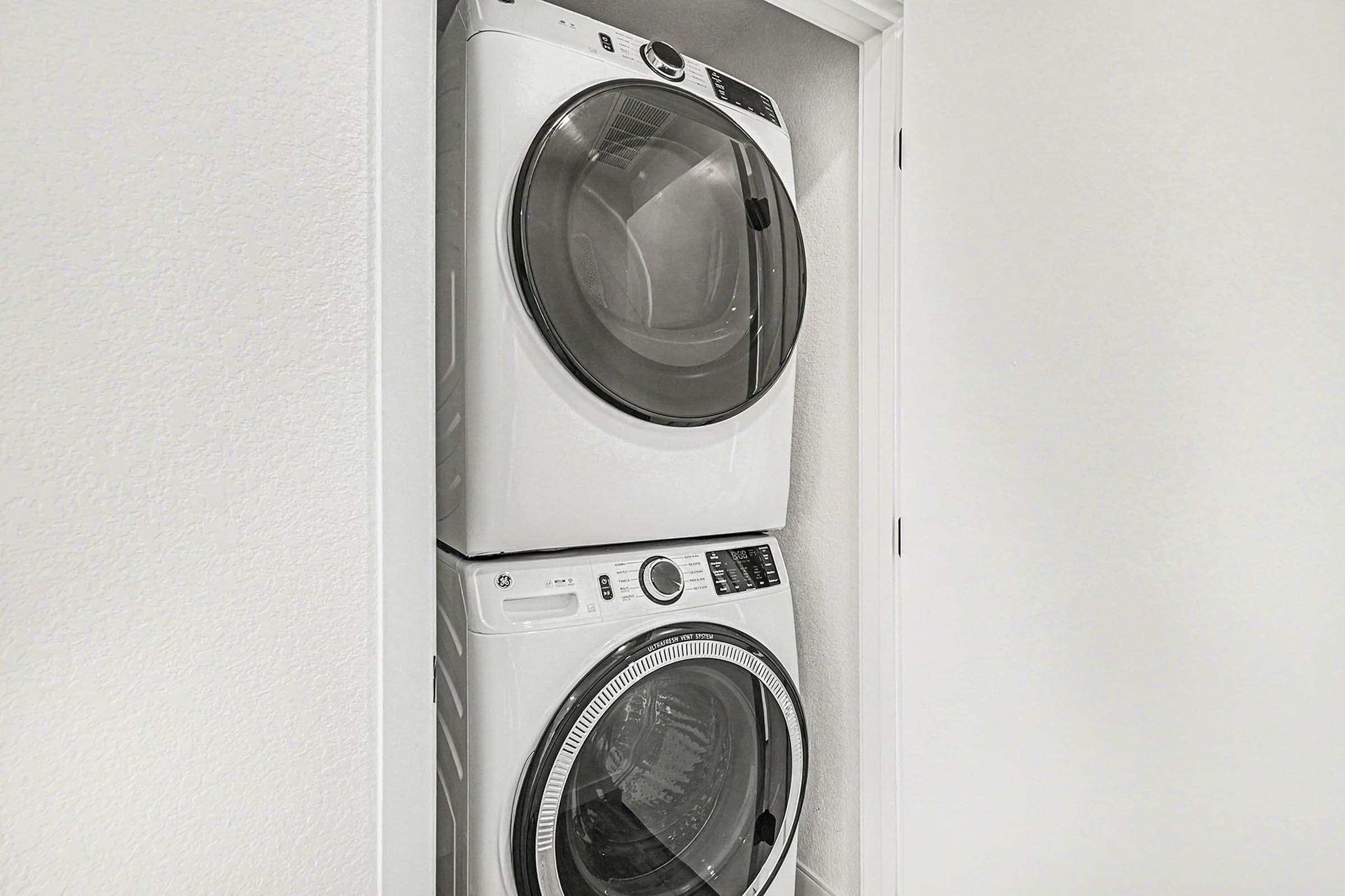 A white stacked laundry washer and dryer set installed in a minimalist laundry space, featuring modern design and clear glass doors, against a neutral wall background.