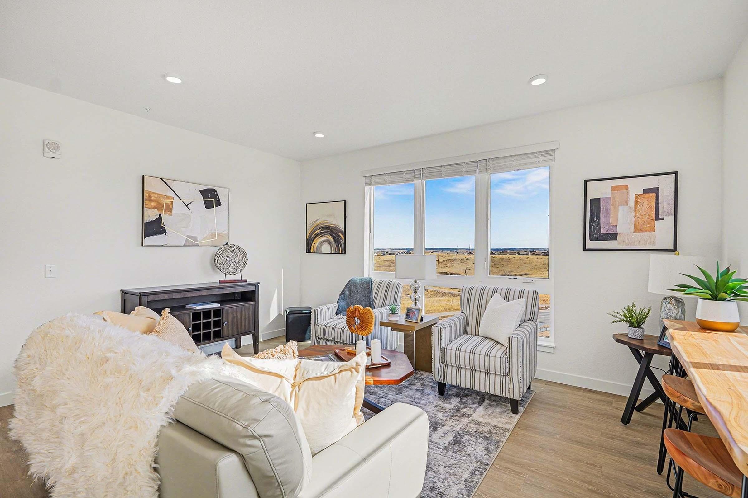 A stylish, modern living room featuring a cozy seating area with a light-colored sofa, patterned armchairs, and a coffee table. There's a large window offering a view of the outdoors, complemented by artwork on the walls and a dining area with wooden chairs and a table.
