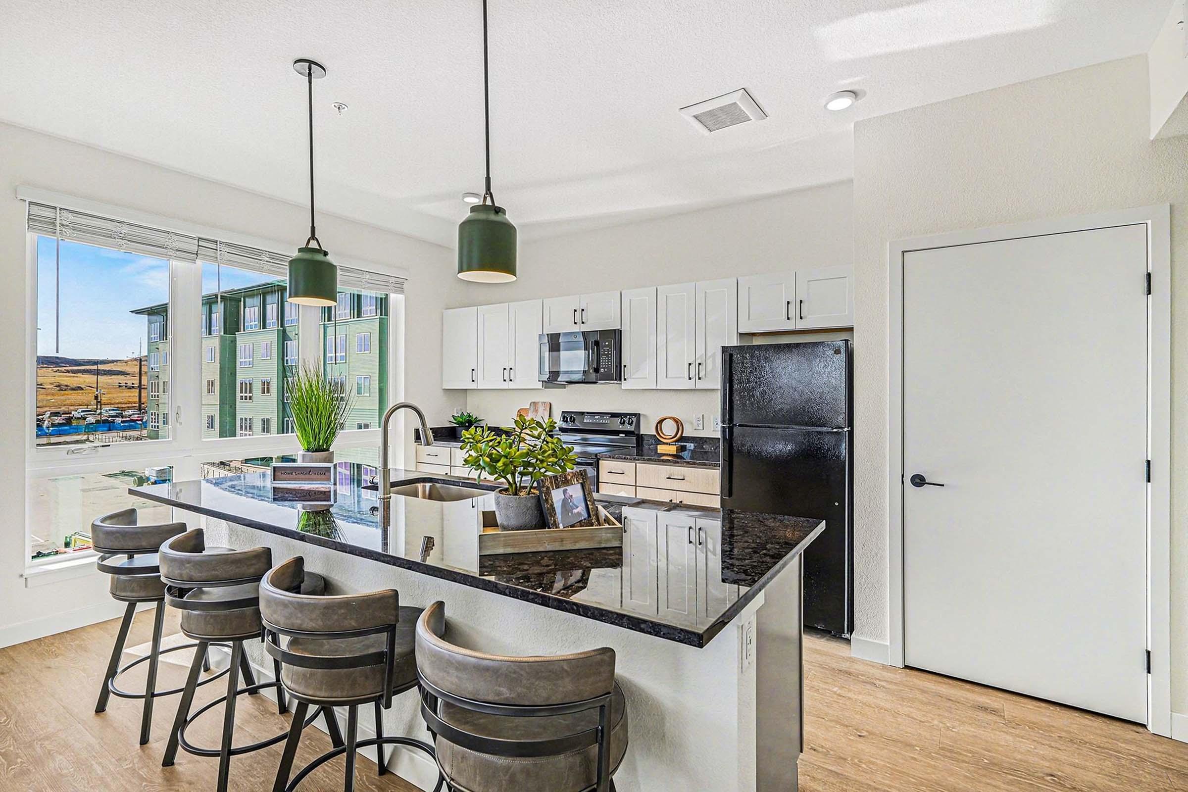 Modern kitchen with a large island featuring bar stools, sleek cabinets, and stainless steel appliances. Bright natural light fills the space from large windows, showcasing a view of surrounding buildings and landscape. Plants add a touch of greenery to the decor.