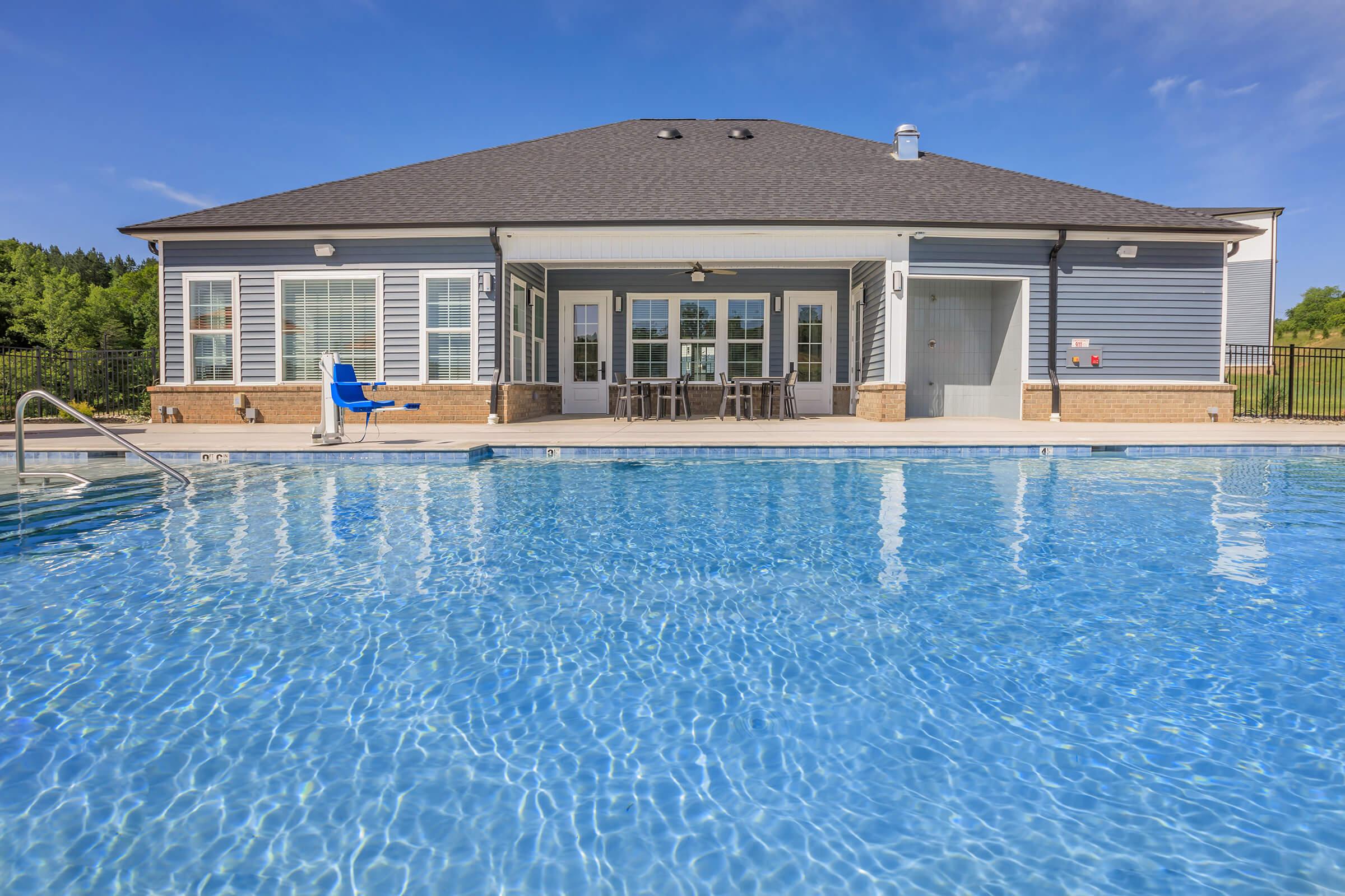A clear blue swimming pool reflects a sunny sky in front of a modern building featuring large windows and a patio area. The pool area includes lounge chairs and a well-maintained landscape, creating a relaxing outdoor environment.