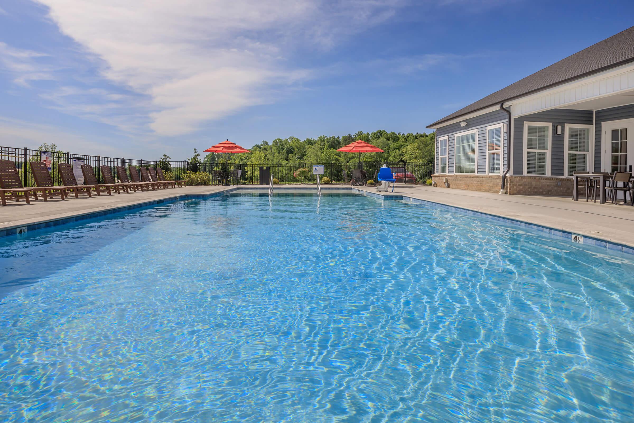 A serene outdoor swimming pool surrounded by lounge chairs and umbrellas. The water is clear and inviting, reflecting a bright blue sky with some clouds. In the background, there's a landscaped area with trees, and a building with large windows adjacent to the pool.