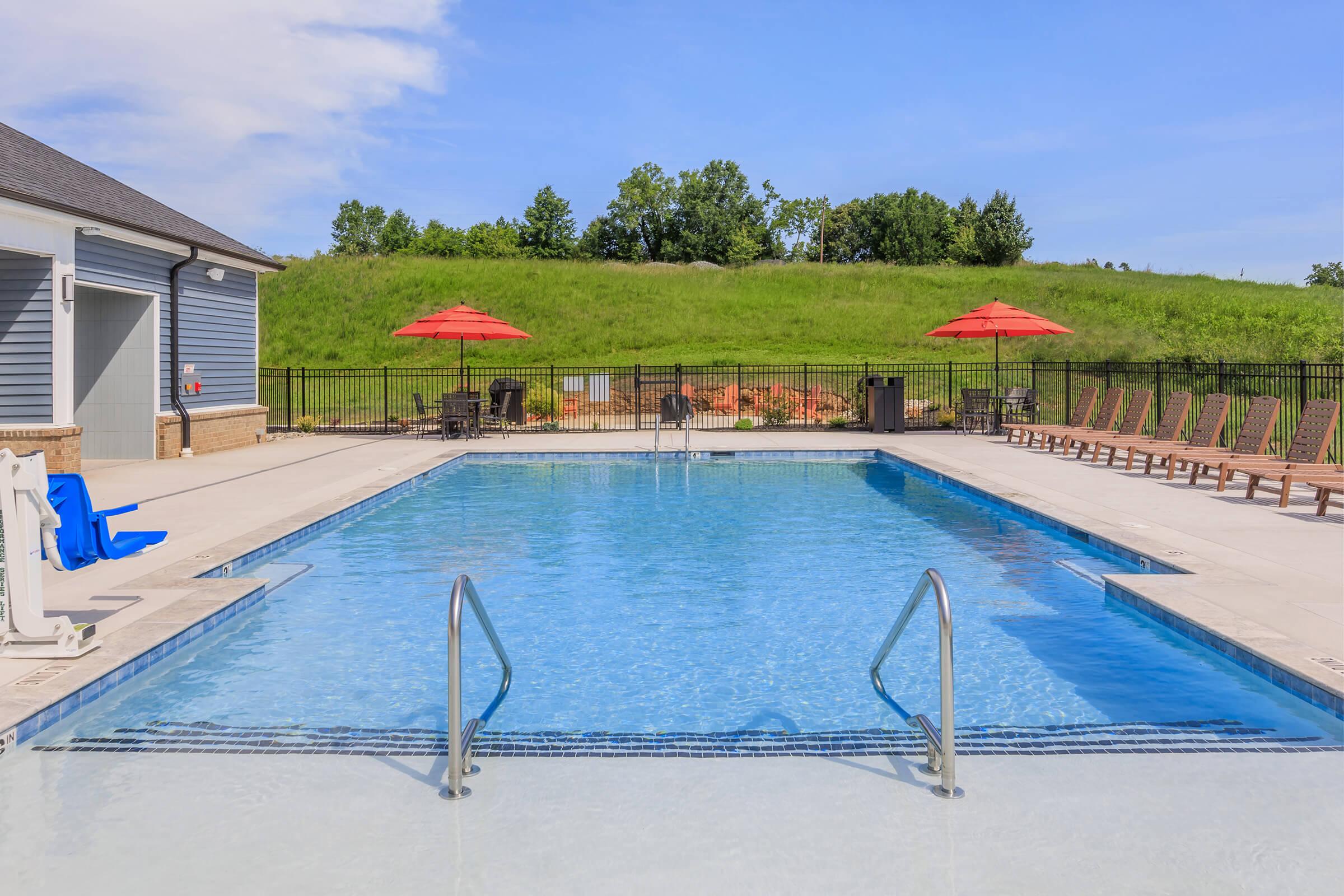 A clear blue swimming pool surrounded by lounge chairs and red umbrellas. In the background, a green grassy hill is visible, along with a fence enclosing the area. Bright blue sky reflects on the water's surface, creating a serene outdoor space for relaxation.