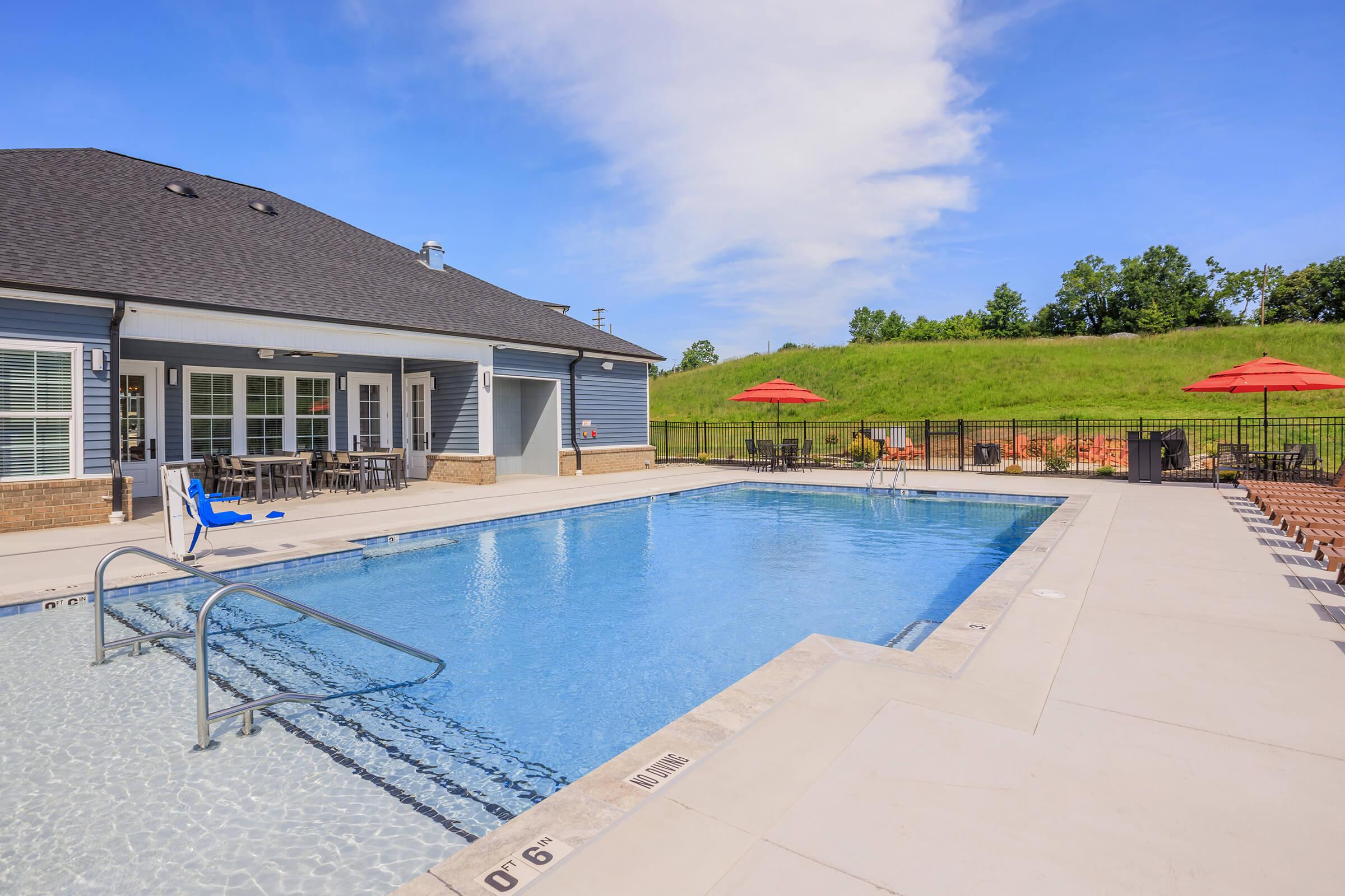 A swimming pool surrounded by a patio area featuring lounge chairs and red umbrellas. In the background, there is a green grassy area with trees. The pool has a ladder for entry and a clear blue sky overhead, creating a relaxed outdoor atmosphere.
