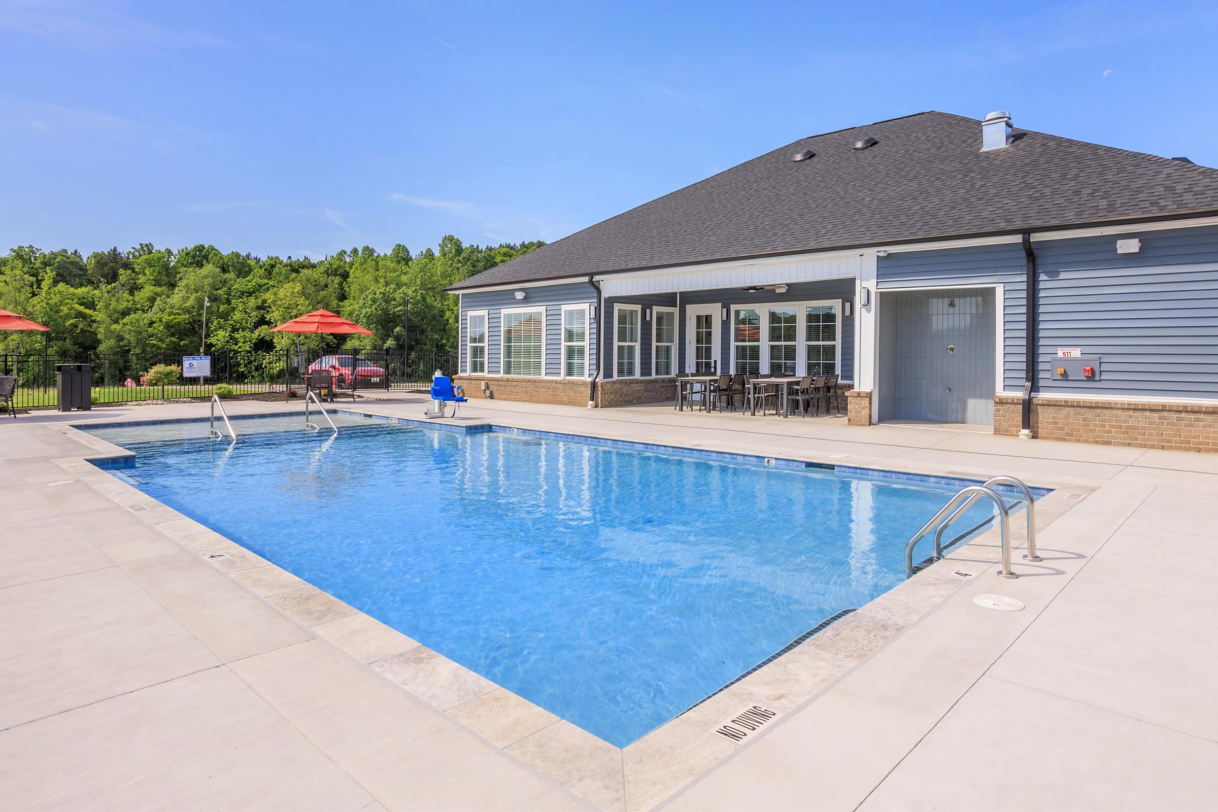 A clear blue swimming pool surrounded by a concrete deck, with two poolside lounge chairs and red umbrellas. In the background, there is a modern building with large windows and tables. Lush green trees line the area, under a bright blue sky.