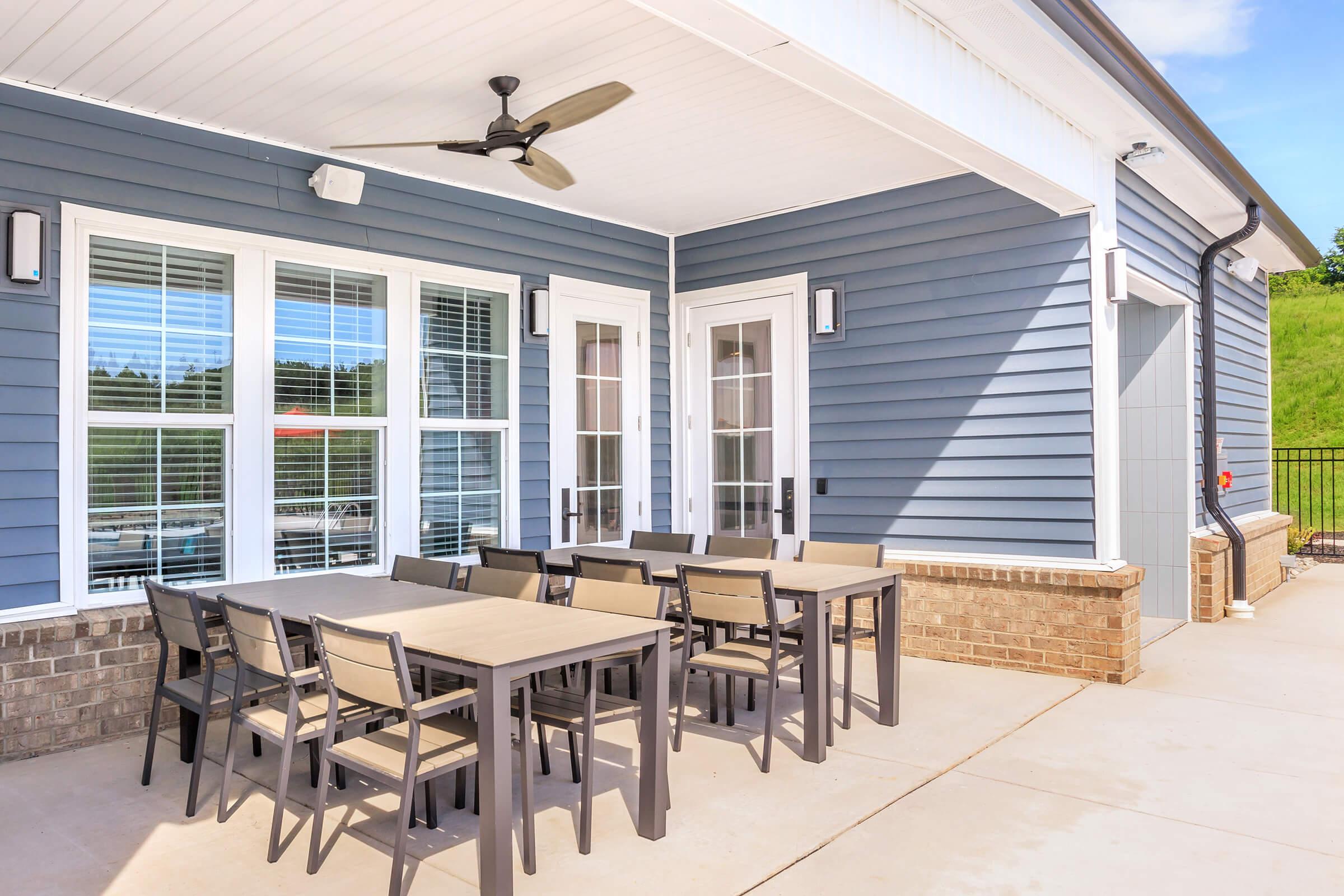 A modern outdoor patio featuring a large rectangular table with multiple chairs, set against a backdrop of blue siding and large windows. The space includes a ceiling fan and is well-lit, creating an inviting atmosphere for dining or gatherings. Green grass is visible in the background.