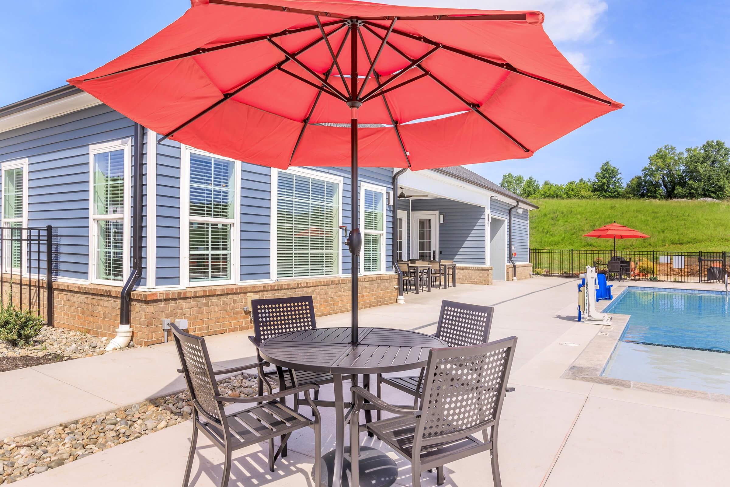 Outdoor patio area featuring a round table with four chairs under a large red umbrella, adjacent to a swimming pool. In the background, a light blue house with large windows and a fenced area. Green grass and trees are visible on the hillside. Bright, sunny day with clear blue skies.