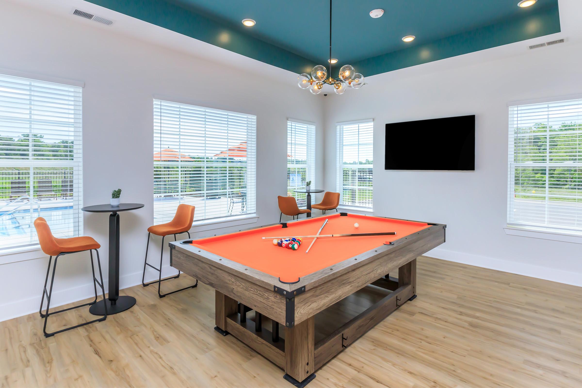 A modern game room featuring a billiards table with an orange felt surface, two orange barstools at a small table, and large windows allowing natural light. A flat-screen TV is mounted on the wall, and the room has a stylish chandelier and a light wood floor. The outdoor view includes greenery.