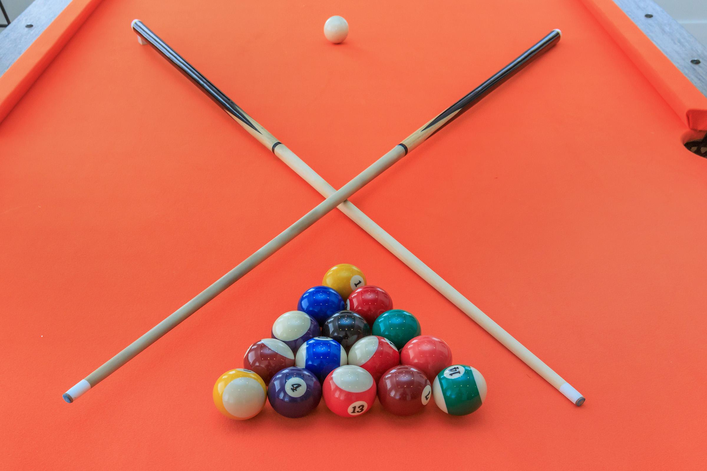 A vibrant orange pool table with two crossed cue sticks and a set of colorful billiard balls arranged in a triangular formation. A single white ball is positioned on the table, ready for a game.