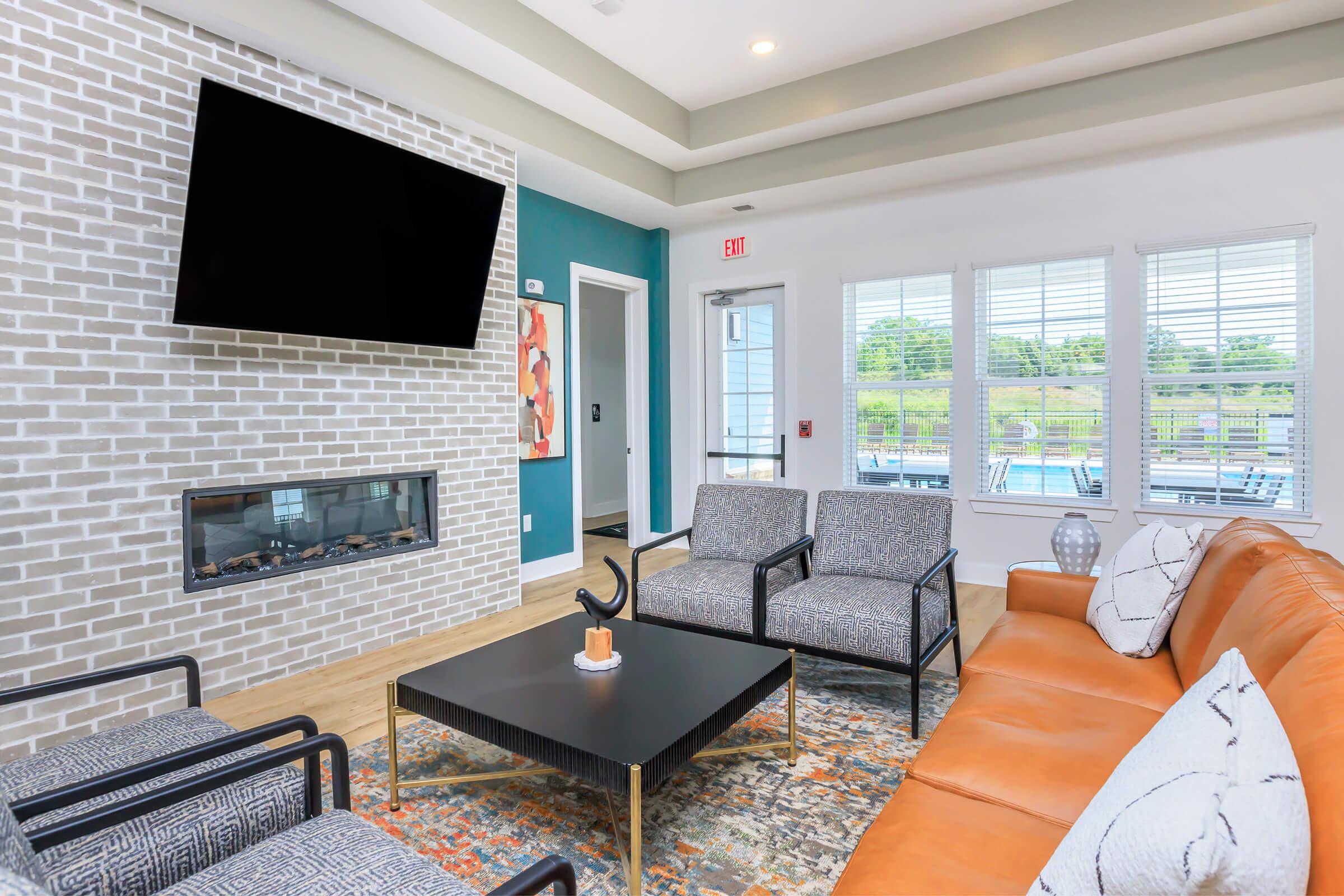 A modern living room featuring a large flat-screen TV mounted on a light brick wall, a cozy orange leather sofa, two stylish armchairs, and a black coffee table. Natural light pours in through large windows, showcasing views of the outdoors. A decorative area rug adds warmth to the space.