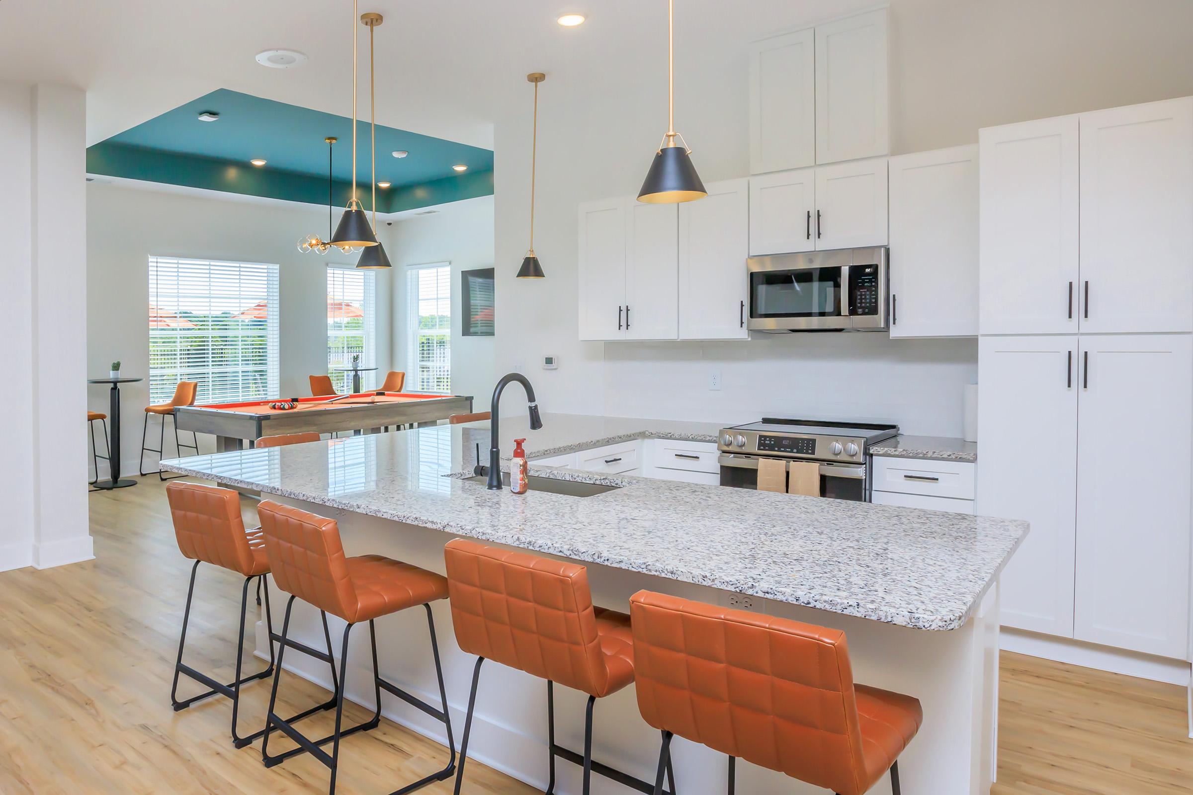 Modern kitchen with a large granite island and four orange bar stools. Stainless steel appliances, including a microwave and oven, are integrated into the white cabinetry. Bright windows allow natural light, and a colorful ceiling adds visual interest. A table for games is visible in the background.