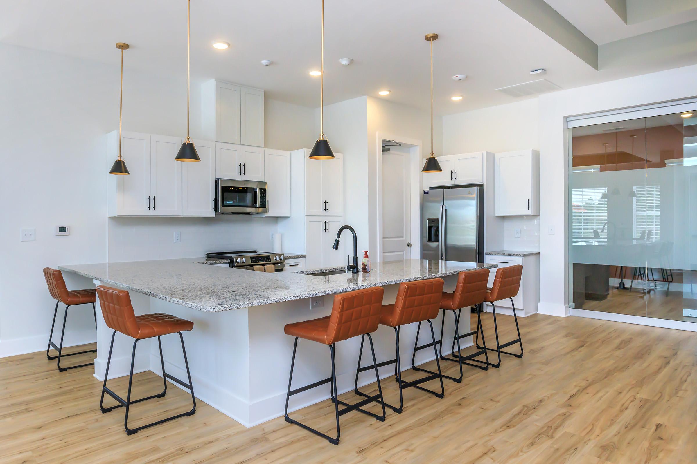 Modern kitchen featuring white cabinets, granite island with seating for eight, stainless steel appliances including a microwave and refrigerator, and pendant lighting. The space has a bright and airy feel with large windows and a wooden floor.