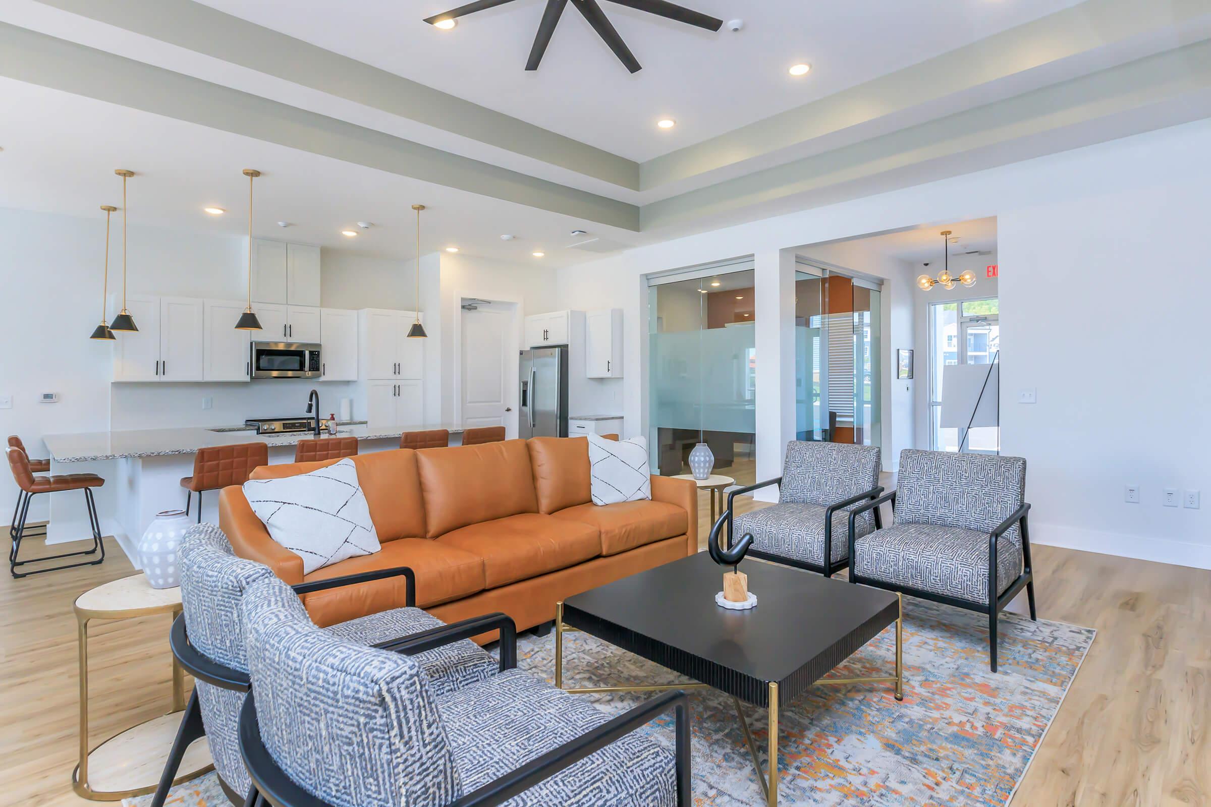 A modern living area featuring a brown leather sofa, two patterned chairs, and a coffee table. The space has a sleek design with light wood flooring, recessed lighting, and large windows. A kitchen area is visible in the background with white cabinets and bar seating.