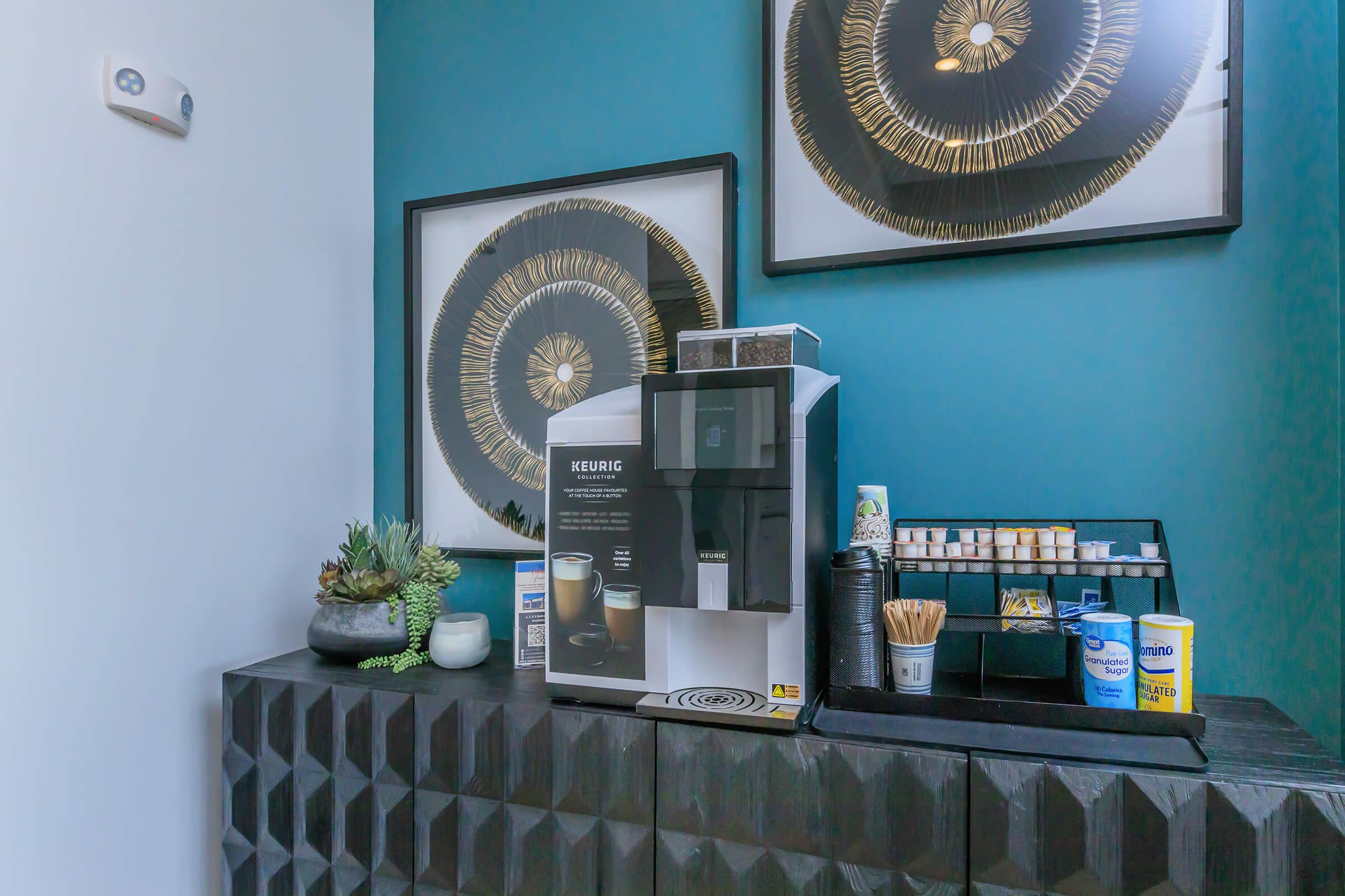 A coffee station featuring a Keurig machine on a textured black cabinet. Behind it, two framed circular wall art pieces are displayed. The station includes various supplies for coffee preparation, such as cups, straws, and wooden stirrers, with some decorative plants for added style.