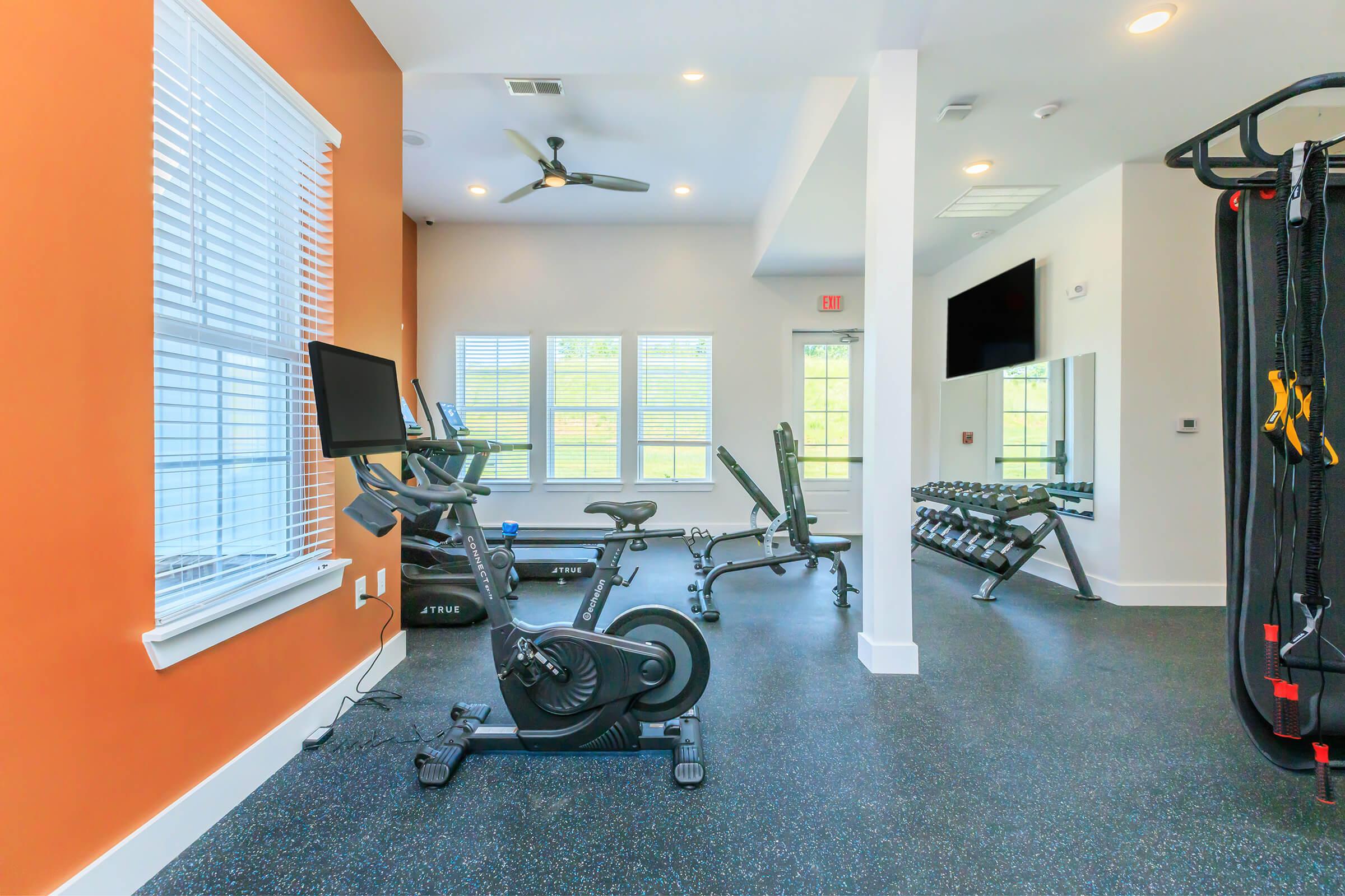A modern fitness room featuring an exercise bike, weight bench, free weights, and a television. The room has large windows allowing natural light, with orange and white walls and rubber flooring for safety. A ceiling fan is installed for ventilation.
