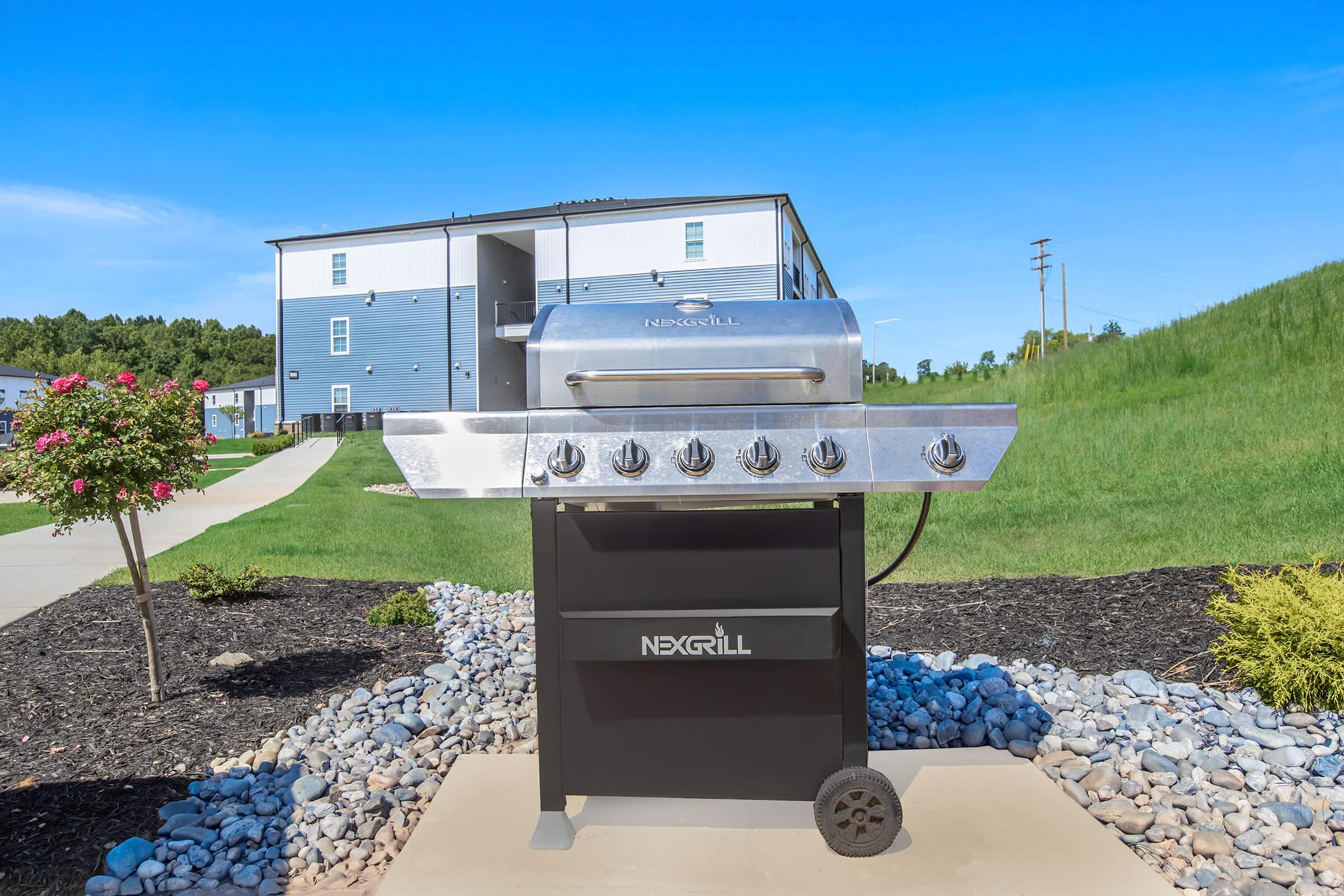 A stainless steel Nexgrill gas grill positioned on a stone path, surrounded by small rocks and landscaped greenery. In the background, a modern building is visible under a clear blue sky, with a well-maintained lawn and flowering plants nearby.