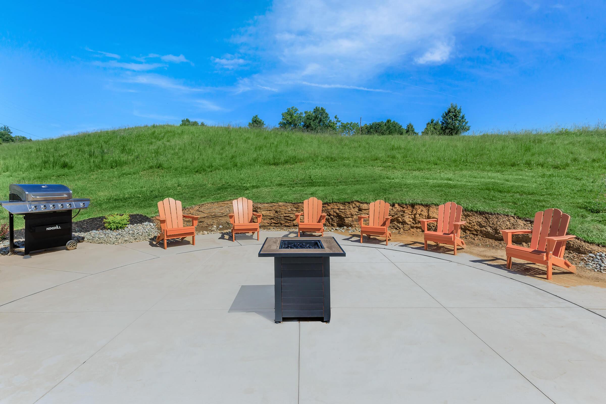 A spacious outdoor patio featuring six orange Adirondack chairs arranged in a semicircle around a black fire pit. A grill sits to the left on a concrete surface, with a lush green hill in the background under a clear blue sky.