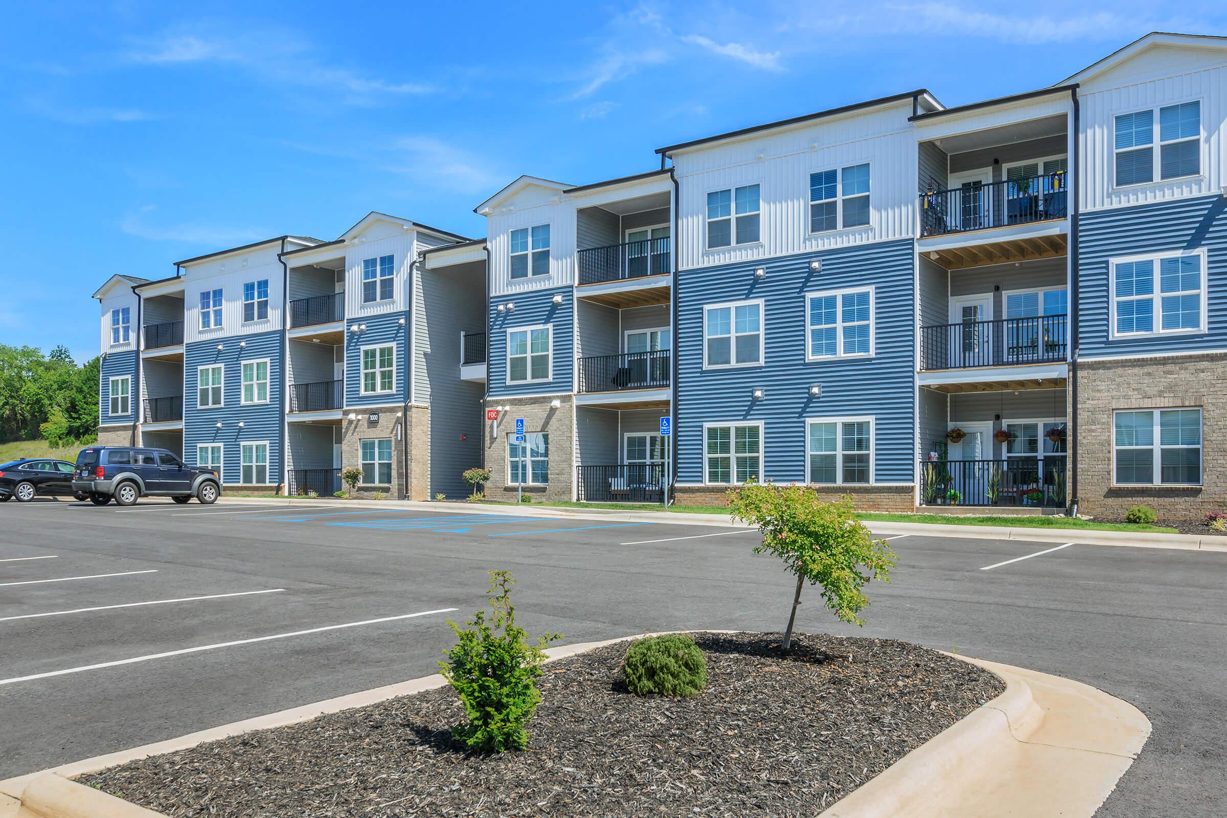 Modern apartment buildings with multiple floors and balconies, situated beside a paved parking lot. Green landscaping with small bushes and mulch in the foreground, under a clear blue sky.
