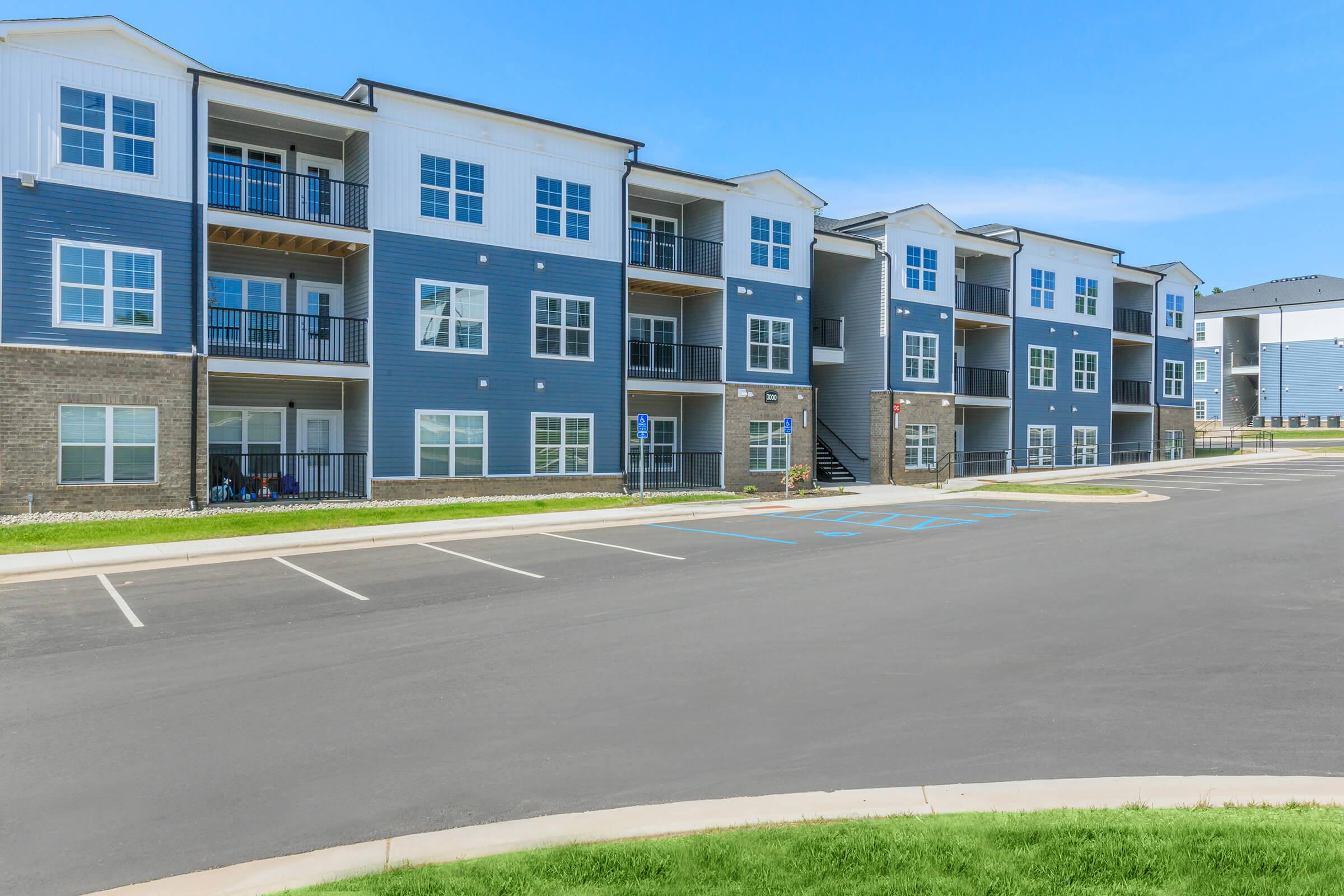 Modern apartment buildings in a residential complex, featuring multiple floors, balconies, and a mix of blue and stone exteriors. The surrounding area includes a paved parking lot and well-maintained green grass, under a clear blue sky.