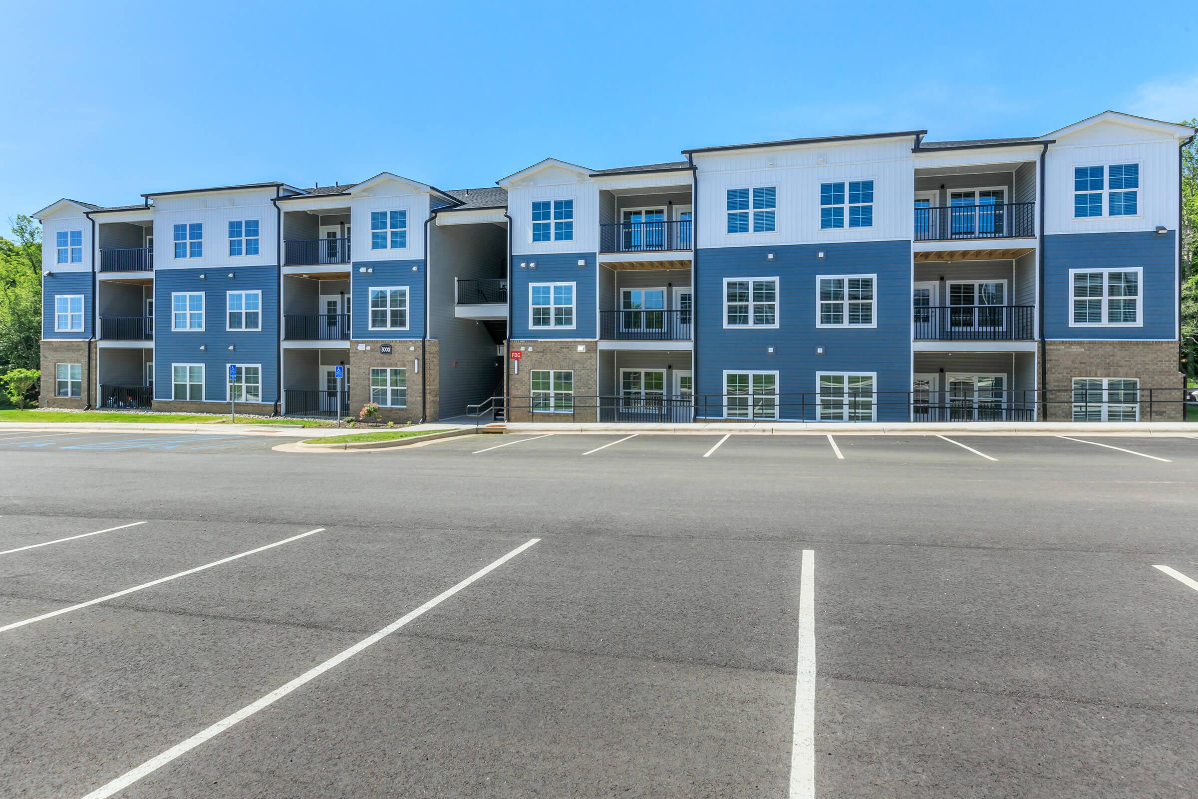 Modern apartment building featuring multiple floors with large windows and balconies. The exterior is a mix of blue and white siding, surrounded by a parking lot with clearly marked spaces. The setting is bright and welcoming, with a clear blue sky in the background.