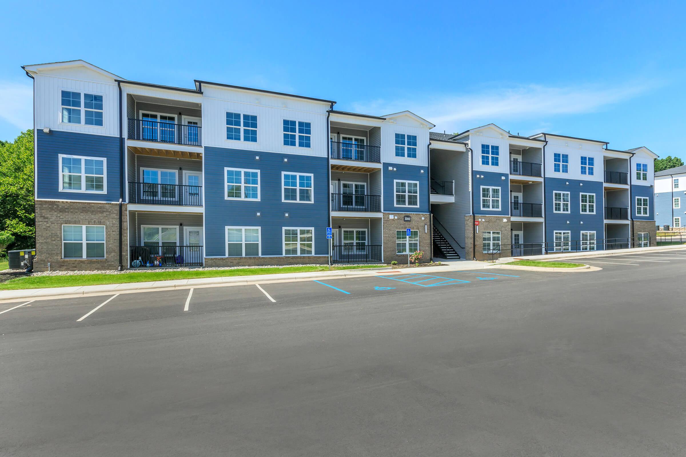 Modern multi-story apartment building with blue and white exterior. The structure features several balconies and large windows. A spacious parking lot is visible in front, with marked parking spaces. Lush greenery surrounds the property under a clear blue sky.