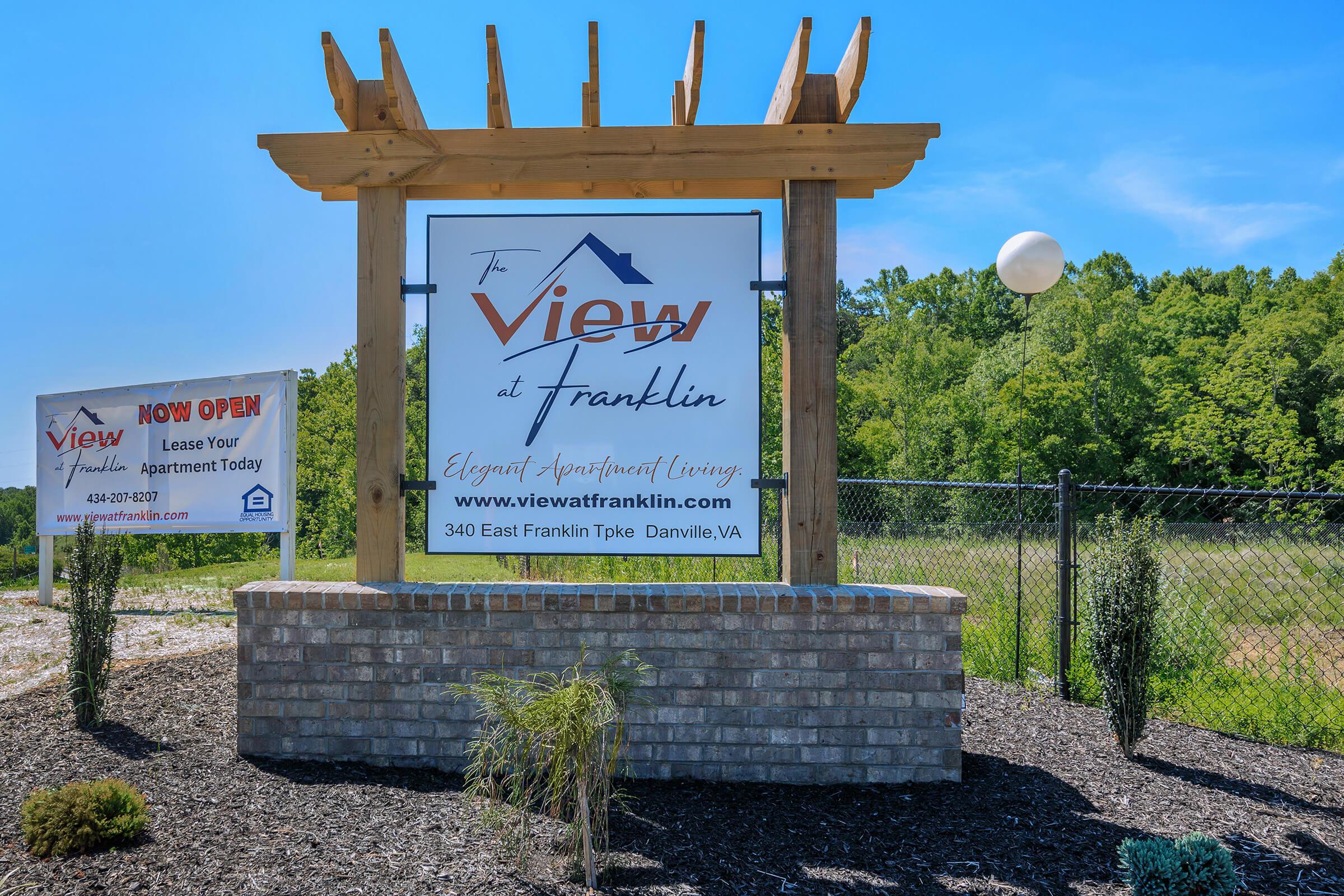 Signage for "The View at Franklin," an apartment community, featuring the slogan "Elegant Apartment Living." The sign is structured with wood framing and a brick base, set against a green landscape with trees. Nearby, another sign indicates availability for leasing apartments.