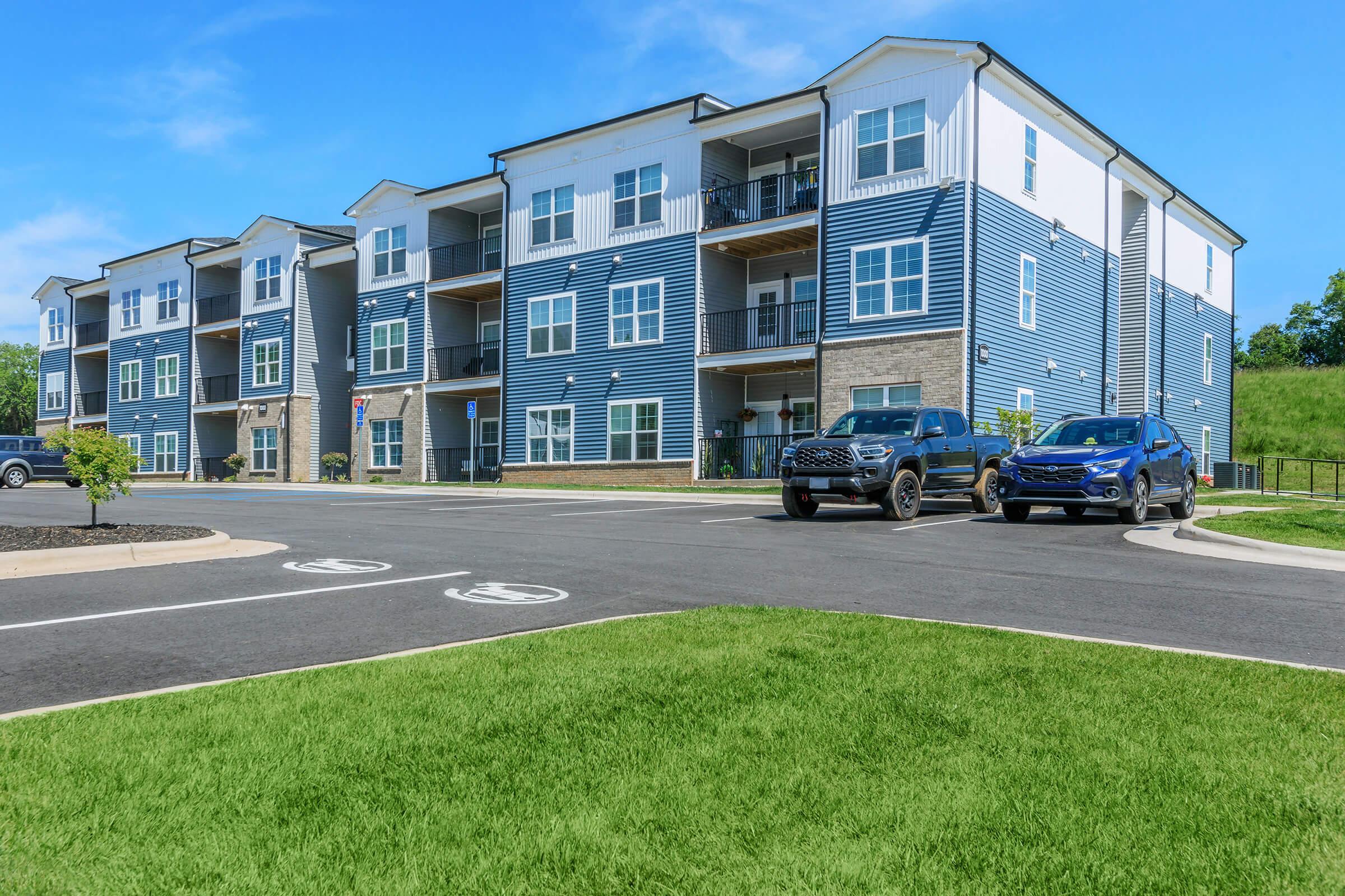 Modern multi-unit apartment building with a mix of blue and gray siding. The structure features multiple balconies and is surrounded by well-maintained landscaping. Two cars are parked in front, with a paved driveway and green grass nearby. Clear blue sky enhances the contemporary look of the complex.