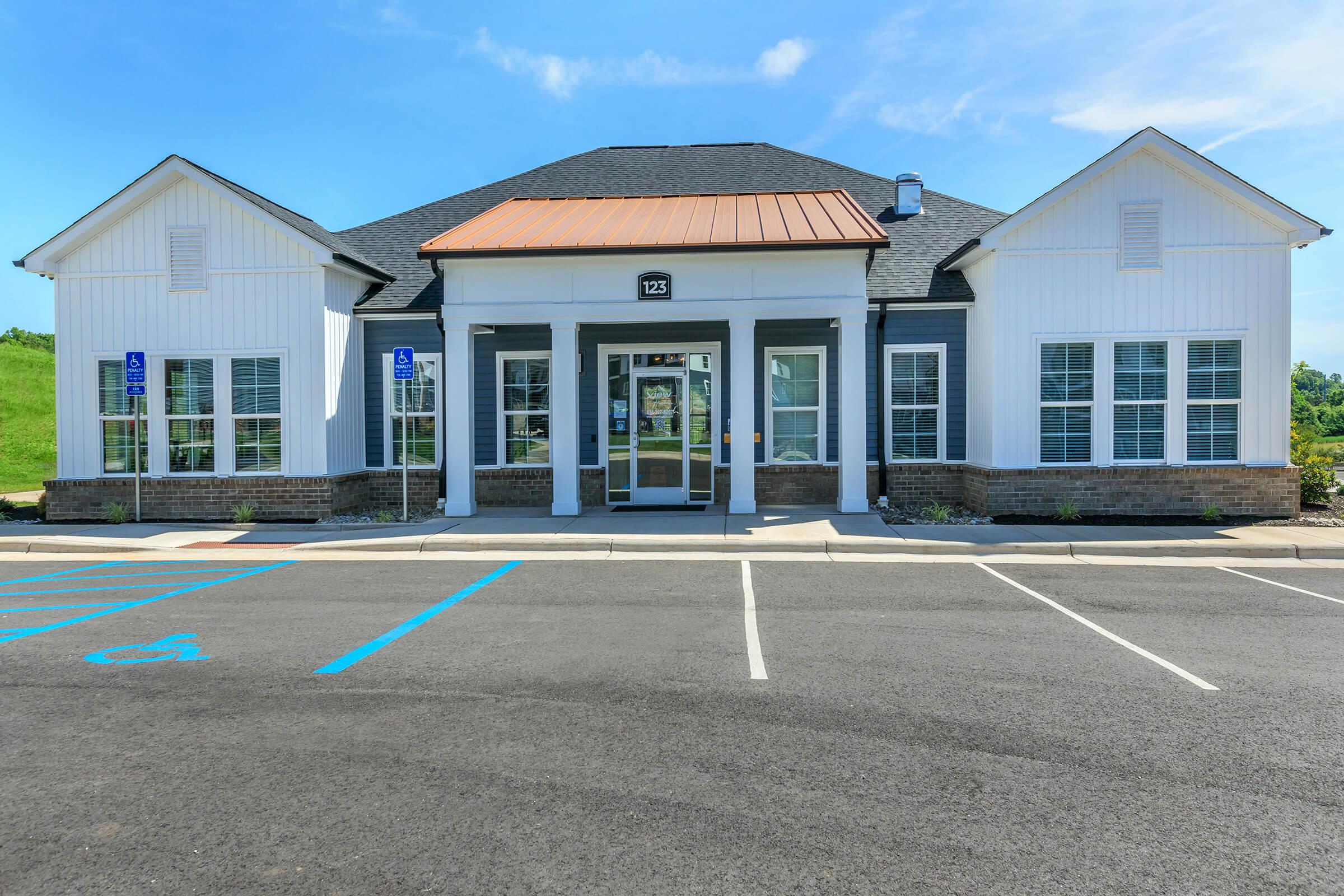 A modern, single-story building with a white exterior and a brown metal roof, featuring large windows and a central entrance. The surrounding area includes a paved parking lot with designated handicap spaces. Green grass is visible in the background under a clear blue sky.