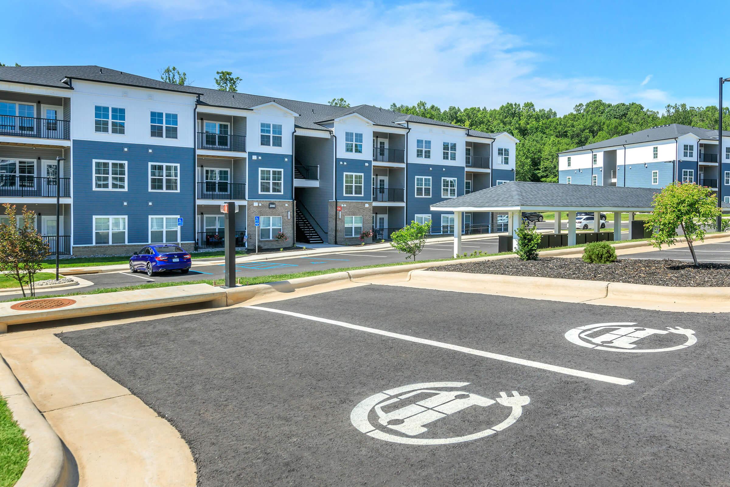 A modern apartment complex featuring multiple two and three-story buildings with gray and white exteriors. In the foreground, a designated electric vehicle parking space is visible, marked with a charging symbol. The scene includes neatly landscaped areas and clear blue skies.