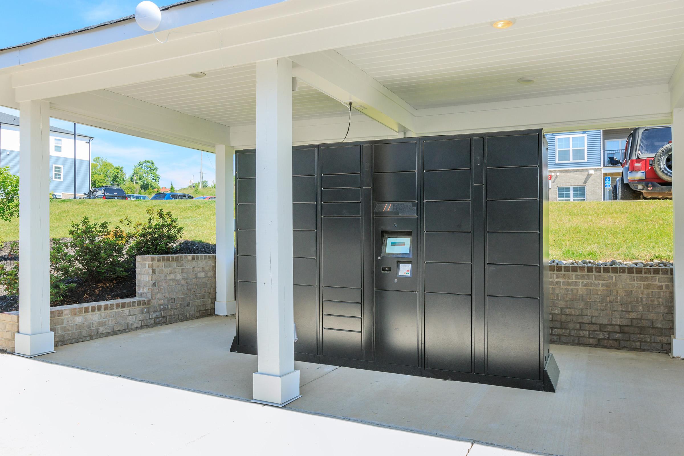 Black package delivery locker with a digital interface, situated under a covered area of a building. Surrounding greenery and a few parked cars in the background, creating a suburban atmosphere. The structure is modern and designed for convenience.
