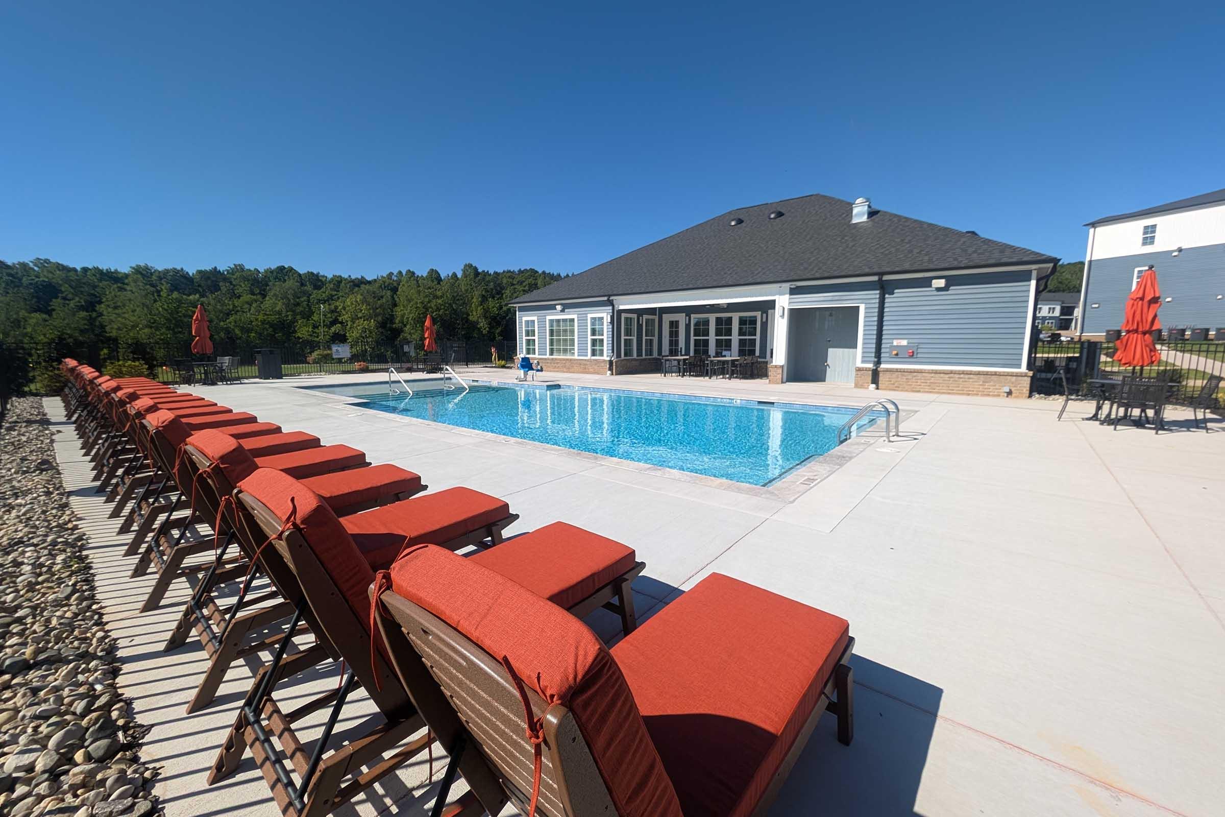 A large outdoor swimming pool surrounded by chaise lounges with orange cushions. Red umbrellas provide shade, and a modern building featuring large windows is visible in the background. Lush greenery lines the area, under a clear blue sky, creating a relaxing atmosphere.