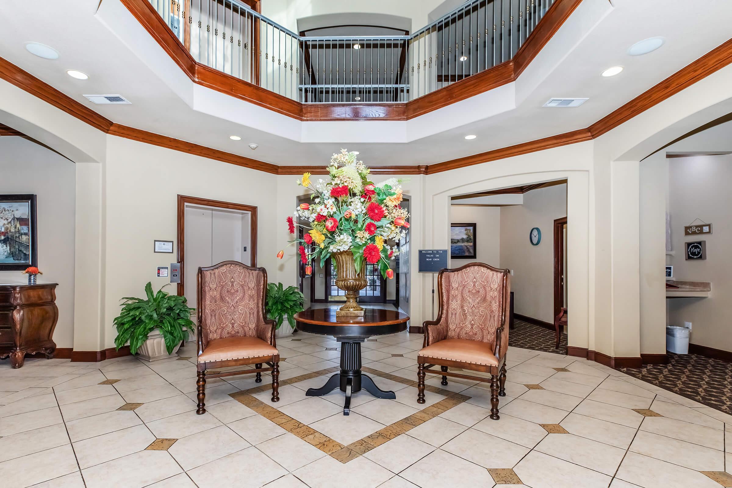 Interior view of a hotel lobby featuring two elegant chairs on either side of a round table with a floral arrangement. The high ceiling adds to the spacious feel, and decorative elements like potted plants and framed artwork enhance the welcoming atmosphere.