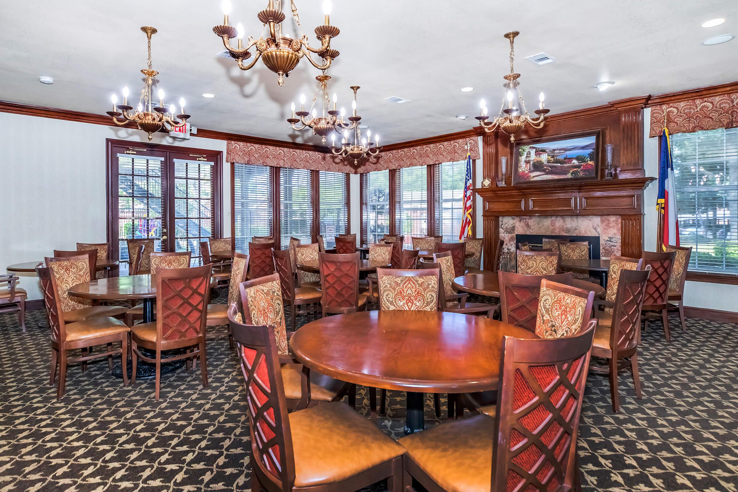 Interior of a dining room featuring multiple round wooden tables and upholstered chairs. The space has elegant chandeliers and large windows, providing natural light. Decor includes rich wood accents and patterned carpets, with a fireplace and framed artwork in the background.