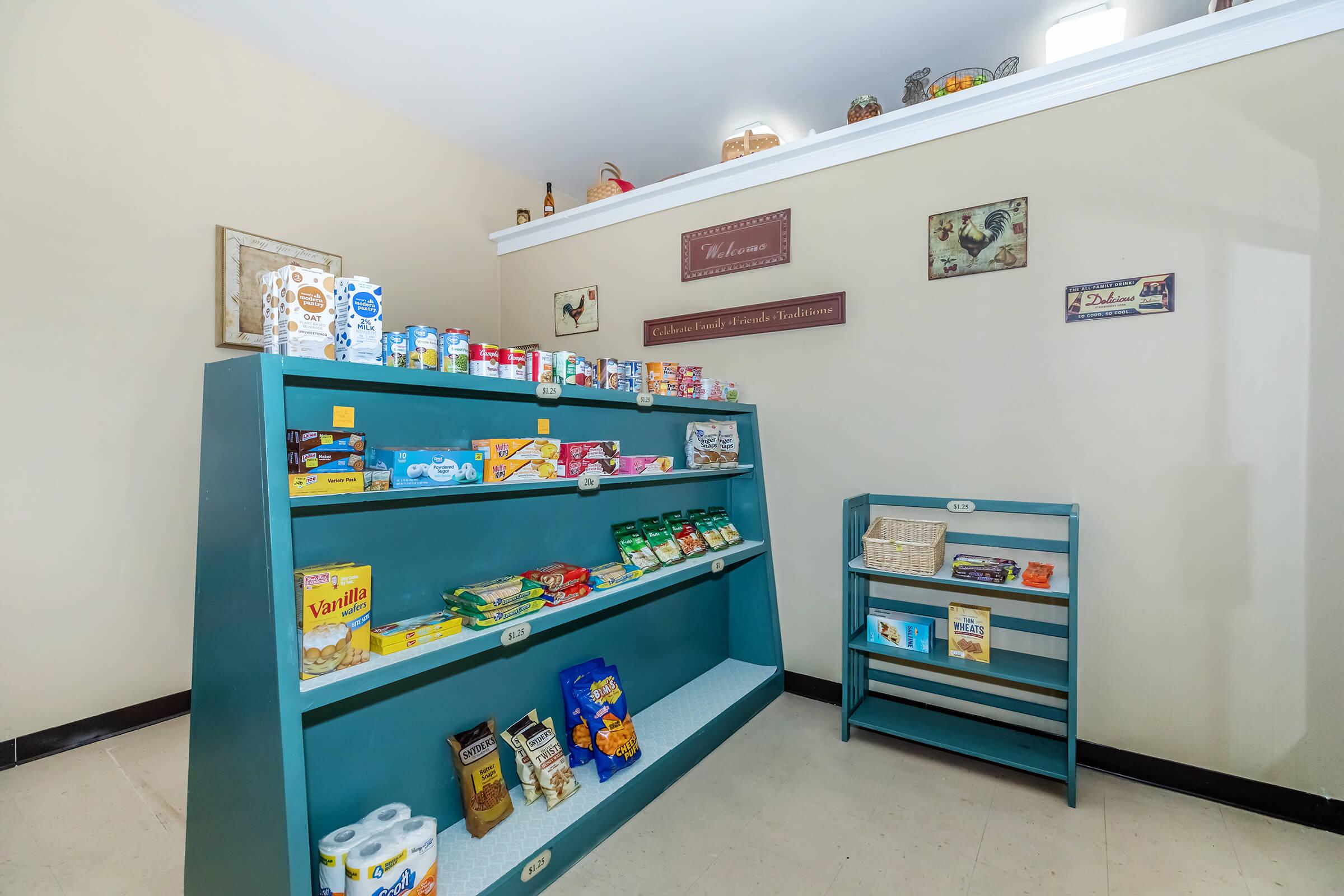 A small market display featuring neatly arranged shelves filled with various packaged food items, snacks, and drinks. The shelves are painted teal and are positioned against a light-colored wall, creating a cozy atmosphere. A few decorative items and signs are placed on the higher ledge above the shelves.
