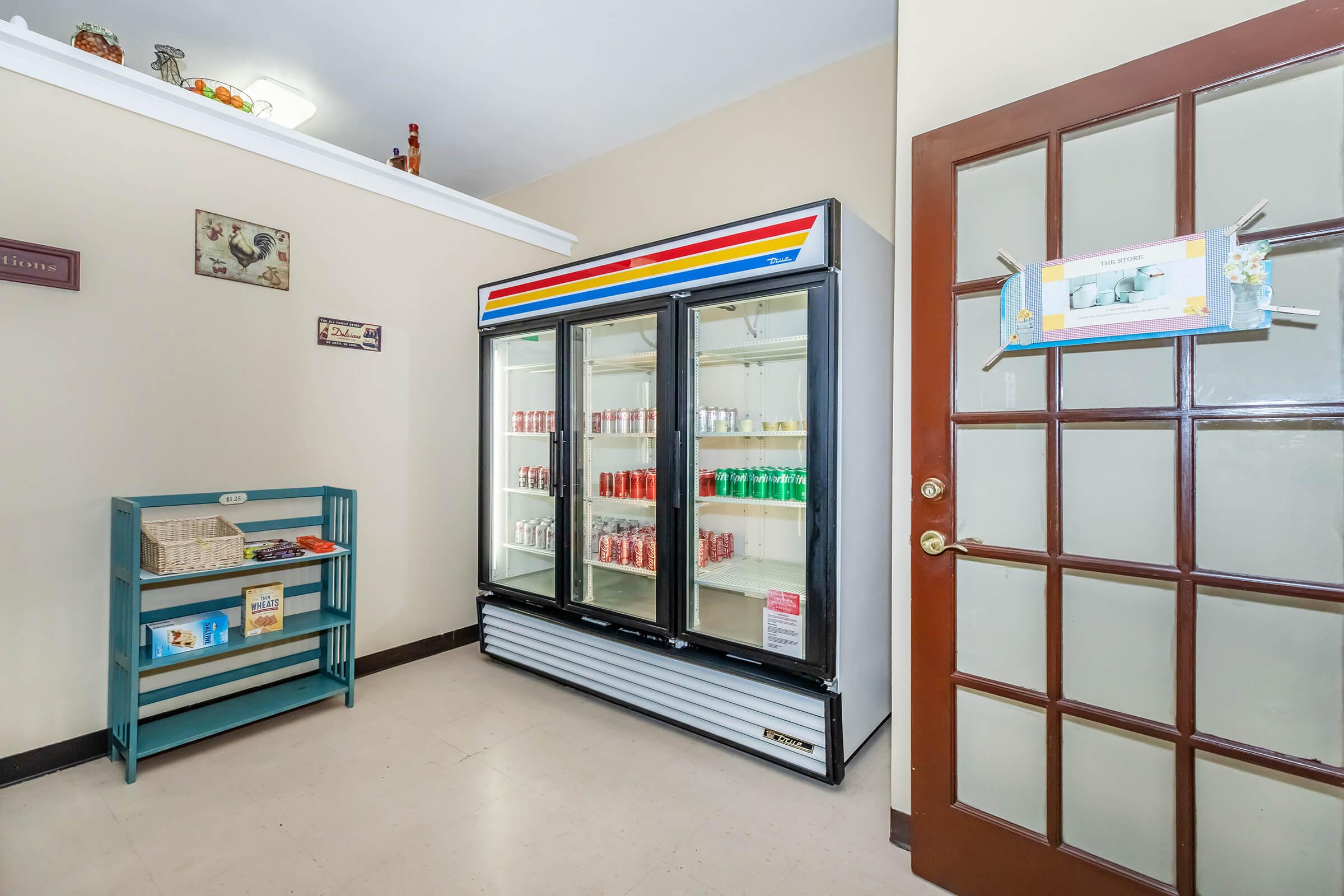 A small room featuring a refrigerator with glass doors displaying various beverages, including sodas. Next to the fridge, there is a blue shelf holding snacks. A door with multiple panels leads outside, and decorative items can be seen on the walls. The overall atmosphere is tidy and inviting.