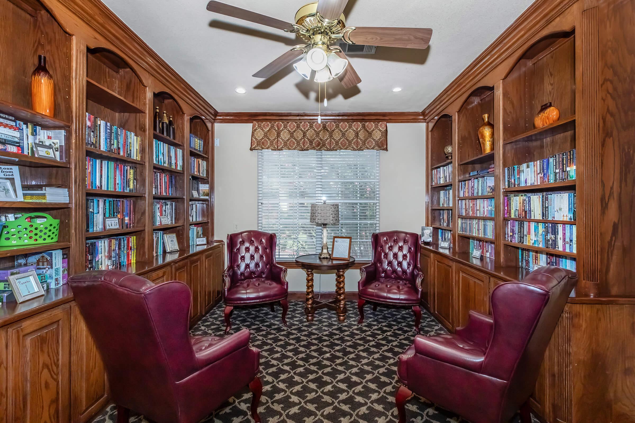 Cozy library corner with two maroon leather armchairs and a small wooden table in between. Bookshelves filled with various books line the walls, and a window with a patterned valance allows natural light to brighten the space. A ceiling fan adds to the welcoming atmosphere.