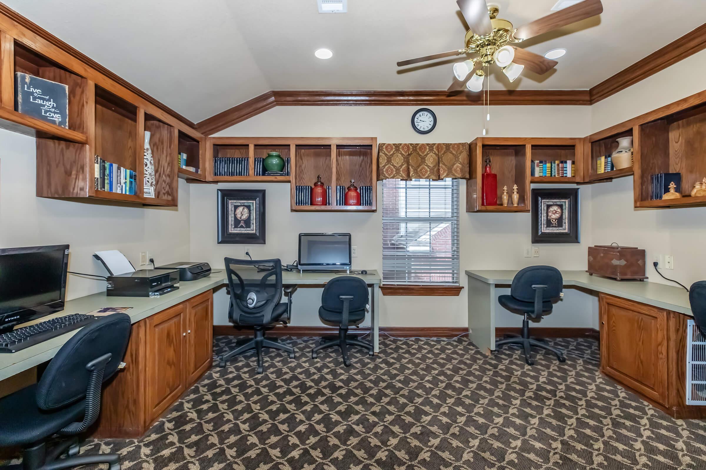 A spacious home office with wooden shelves featuring decorative items and books. Two workstations with black chairs are equipped with computers. The room has a ceiling fan, a wall clock, and a window covered with patterned curtains, providing a cozy and organized workspace atmosphere.