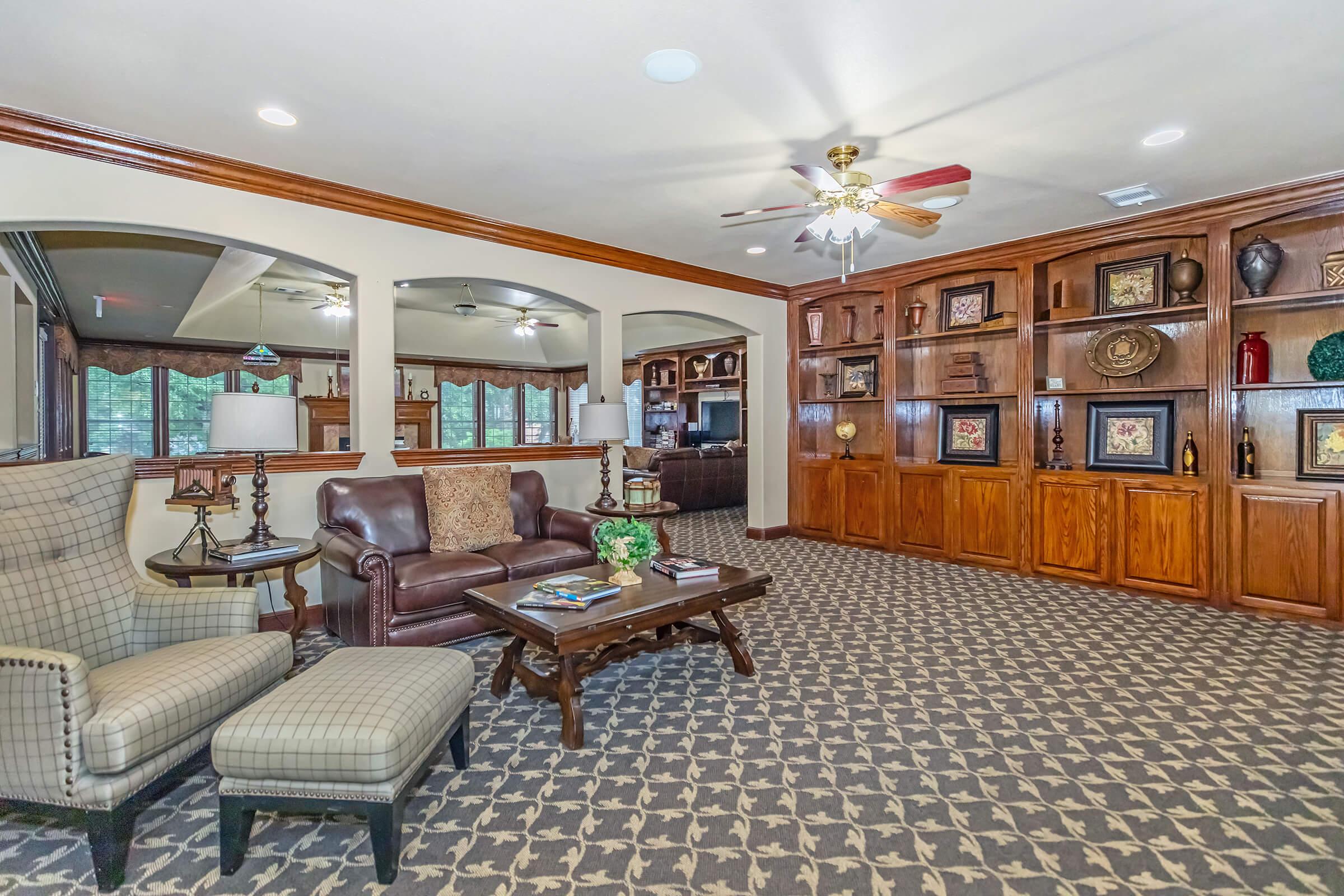 A cozy living area featuring a brown leather sofa, an armchair with a checkered pattern, and a wooden coffee table. The walls have built-in shelves with decorative items. Large windows provide natural light, and the carpet has a geometric pattern, creating an inviting atmosphere.