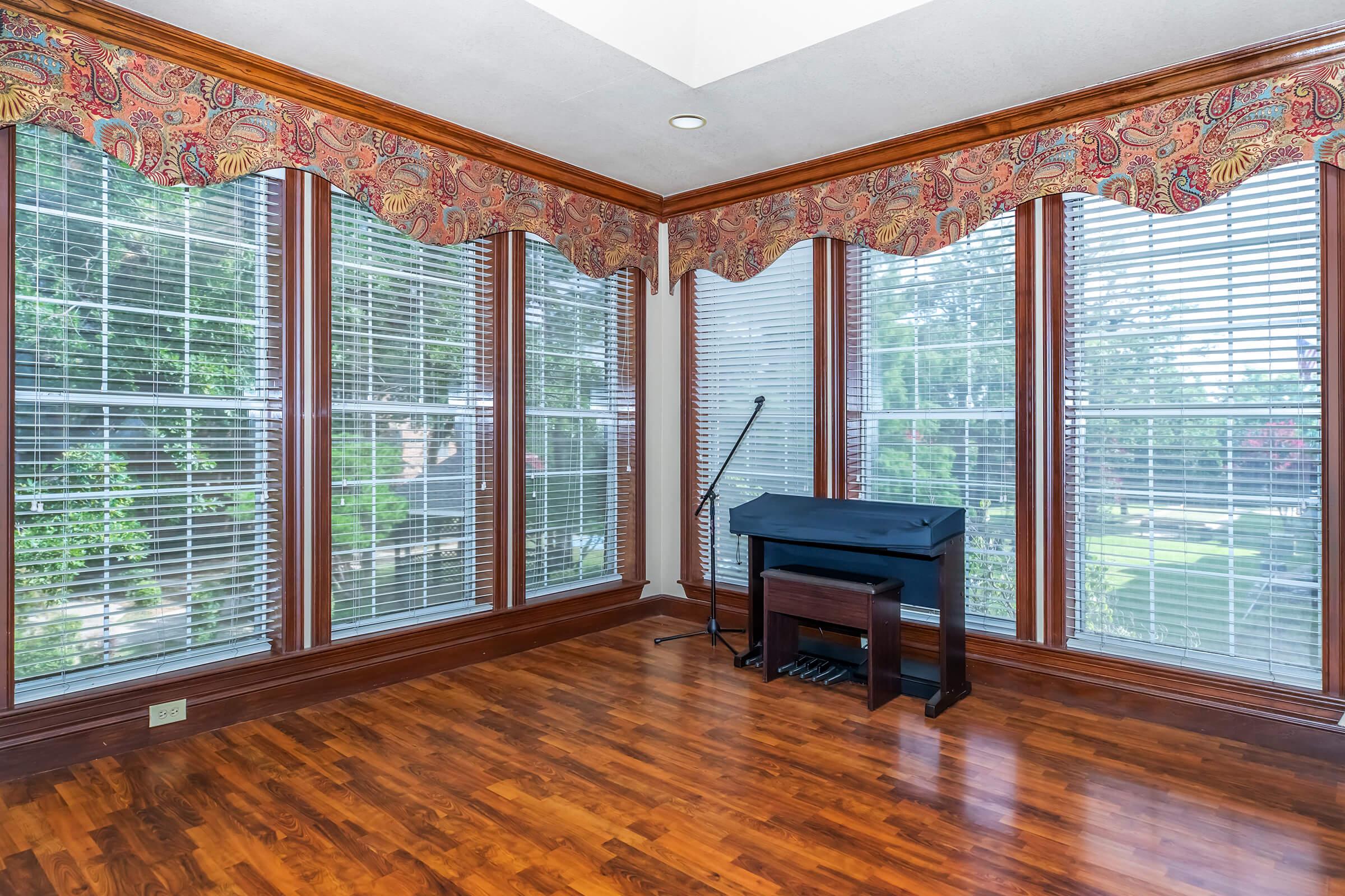 A bright room with large windows featuring wooden frames and blinds, adorned with patterned valances. The floor is polished hardwood, and there is a black piano positioned near the windows, with a music stand beside it, creating a warm and inviting atmosphere.
