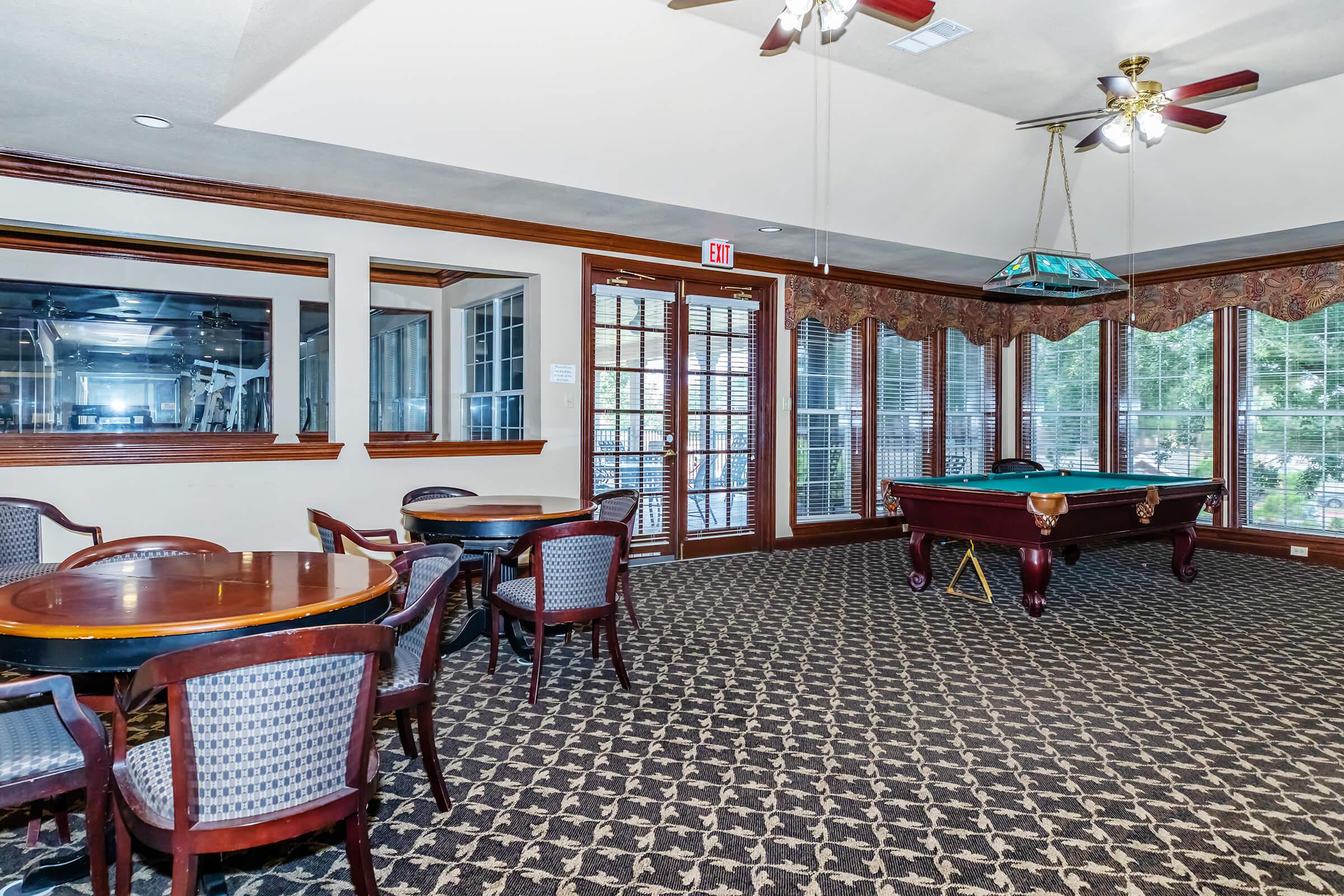 An elegantly furnished game room featuring a pool table, circular and rectangular dining tables with patterned chairs, and large windows allowing natural light. The decor includes wooden accents and a stylish ceiling fan. Subtle lighting enhances the inviting atmosphere.