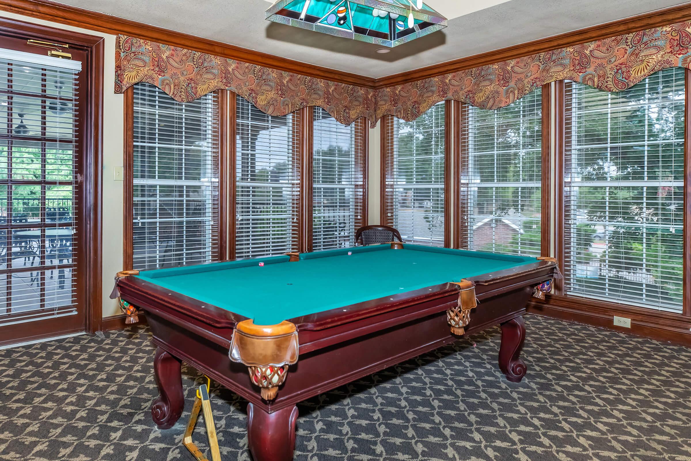 A well-lit game room featuring a green pool table with ornate wooden details, surrounded by large windows with blinds. The room has a patterned carpet and decorative curtains, creating a cozy atmosphere for playing billiards.