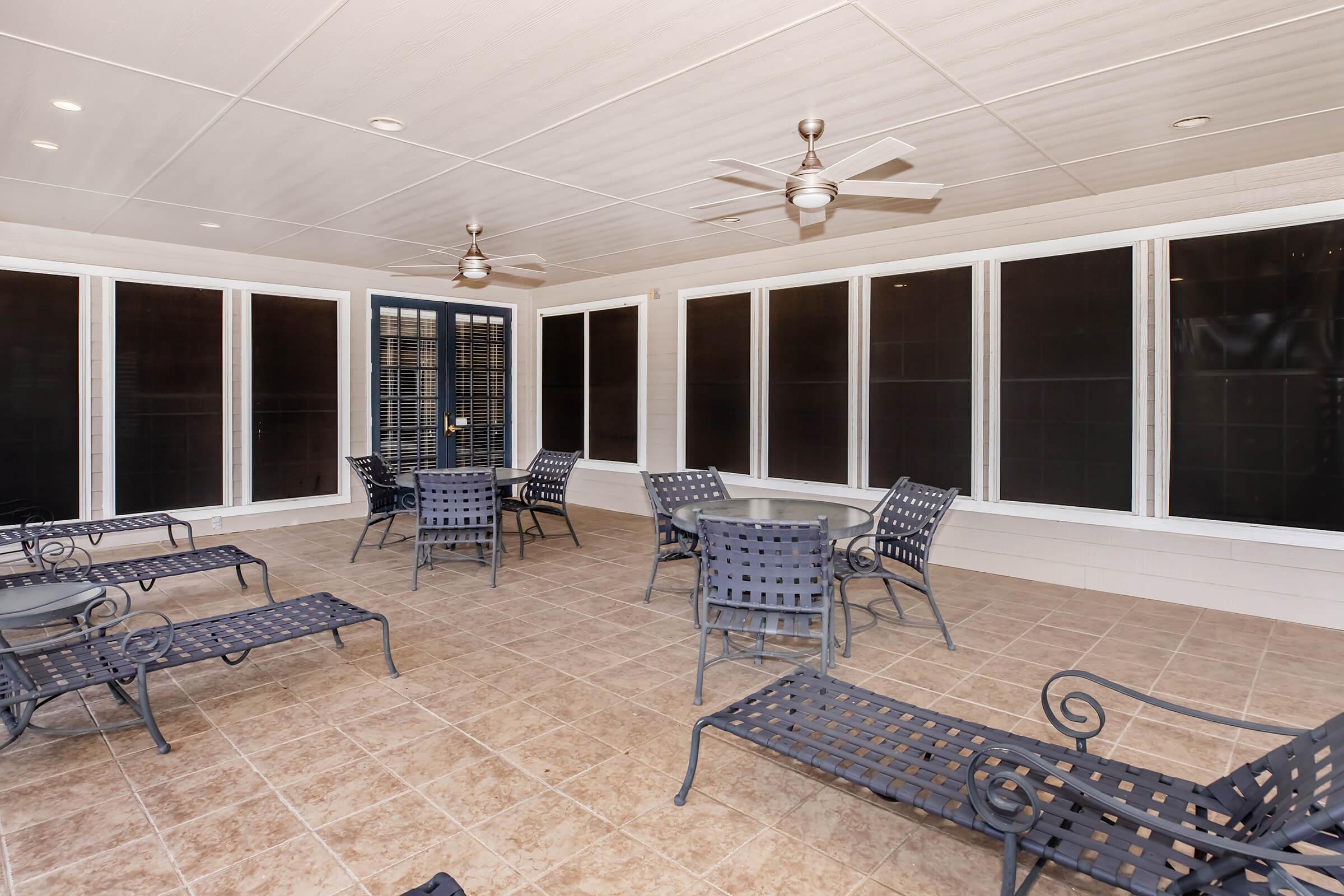 A covered patio area featuring several metal lounge chairs and tables. Large windows allow for natural light, and ceiling fans are installed for ventilation. The floor is tiled, and the overall space is designed for relaxation and socializing.