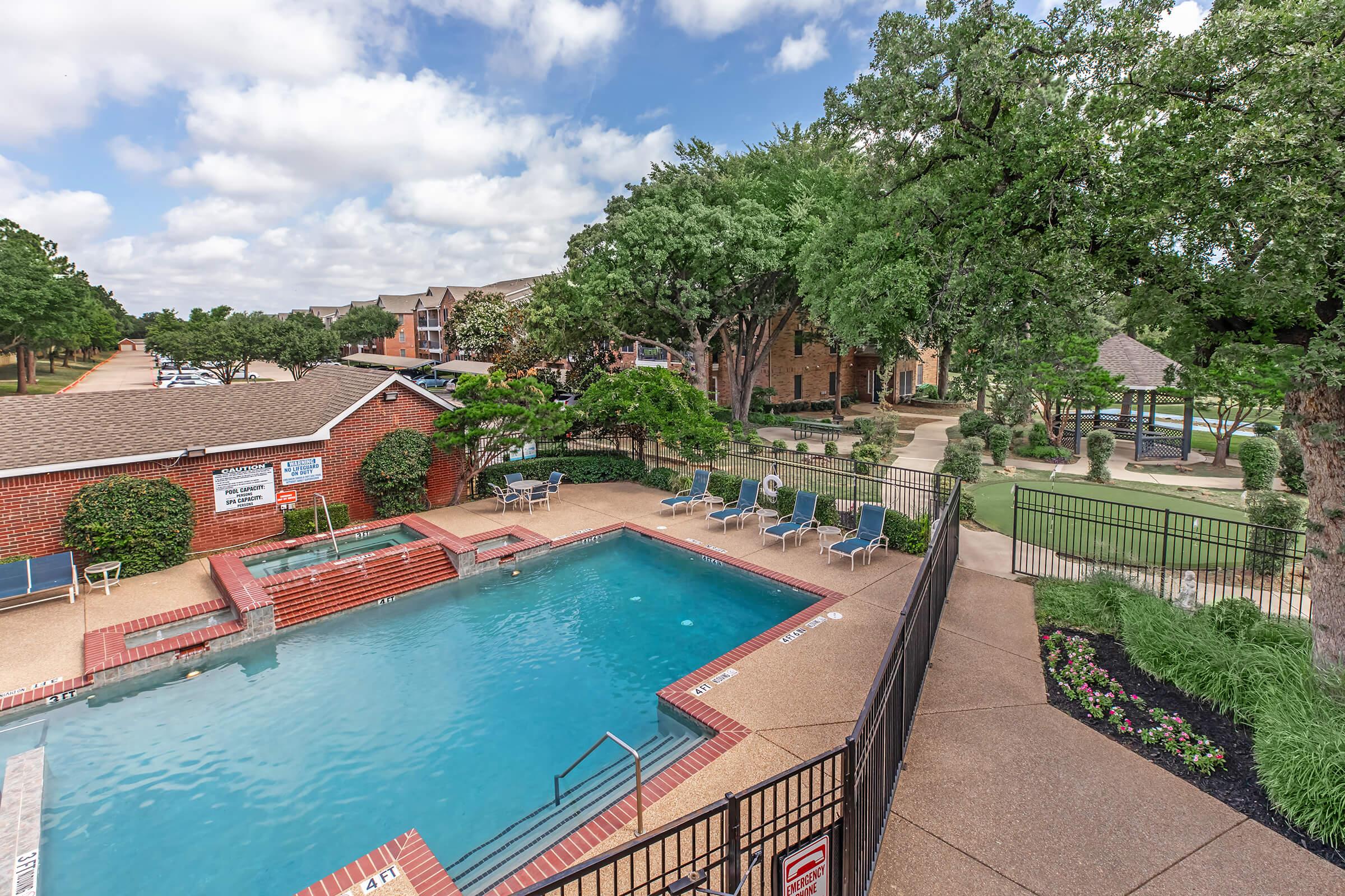 A view of a swimming pool surrounded by lounge chairs and landscaping. The pool area features steps leading into the water. Nearby, there are tree-lined pathways and well-kept greenery. Apartments are visible in the background, creating a relaxing residential atmosphere.