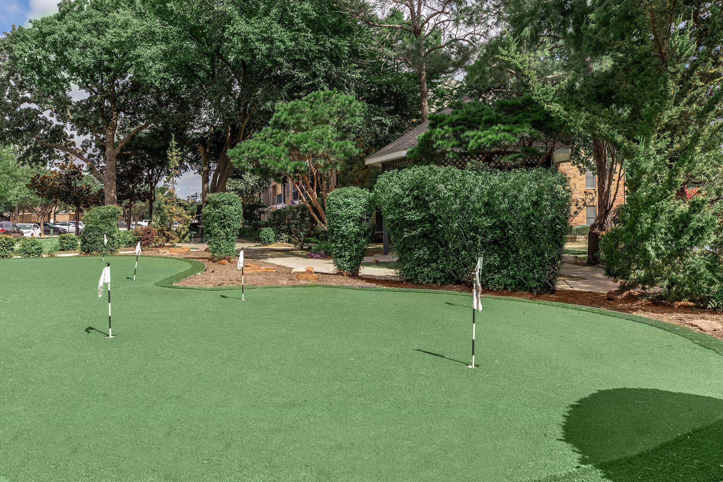 A well-maintained putting green featuring several white flags, surrounded by lush greenery and bushes. In the background, a building can be seen among the trees, creating a serene outdoor setting ideal for golf practice or leisure activities.