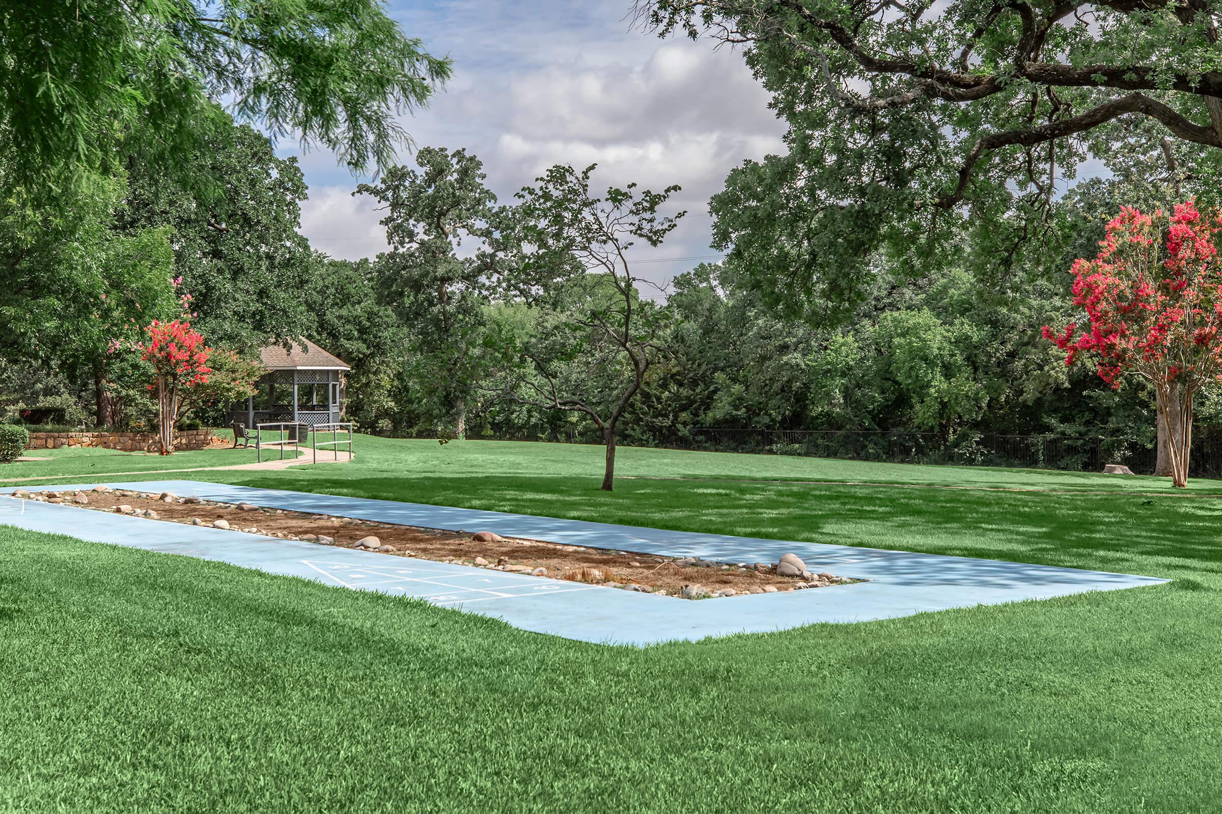 A spacious green lawn featuring a rectangular area outlined in blue, with a dirt patch in the center. Trees and shrubs surround the area, and a small gazebo is visible in the background under a cloudy sky. Brightly colored trees add a splash of color to the landscape.