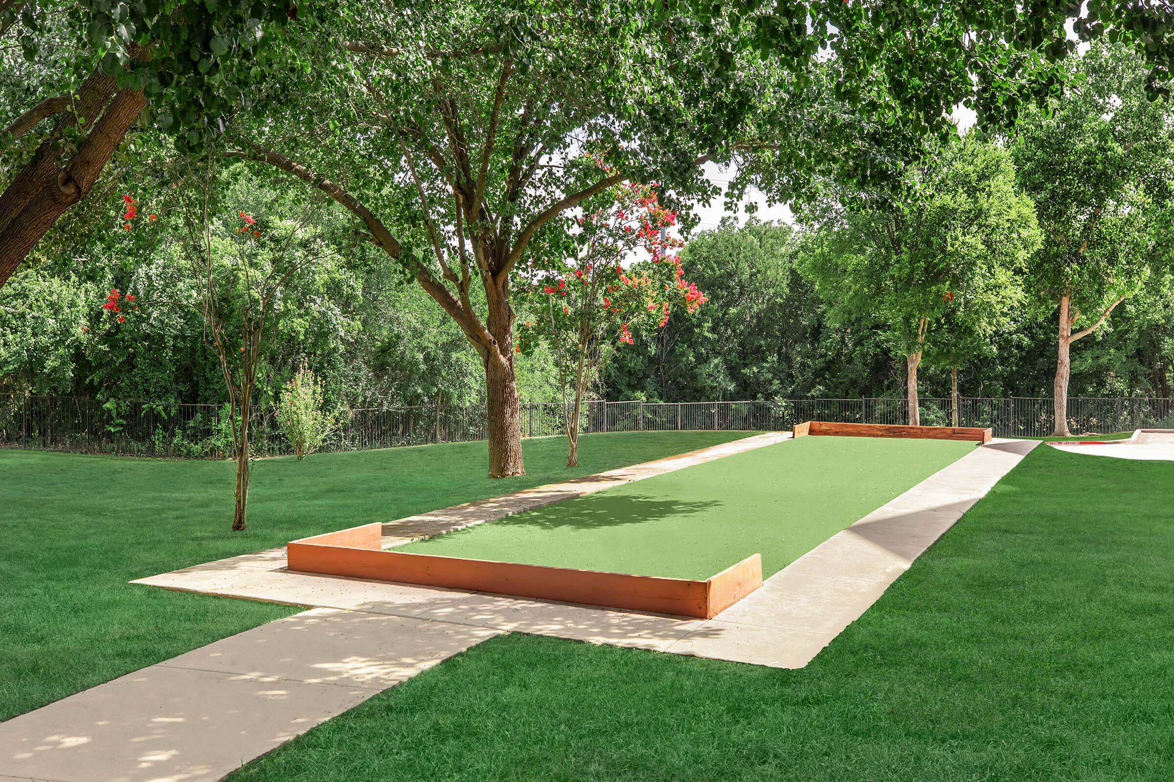 A well-maintained outdoor bocce ball court surrounded by lush green grass and trees. The court features a smooth surface with wooden borders and is set in a tranquil park-like setting, ideal for recreational play.