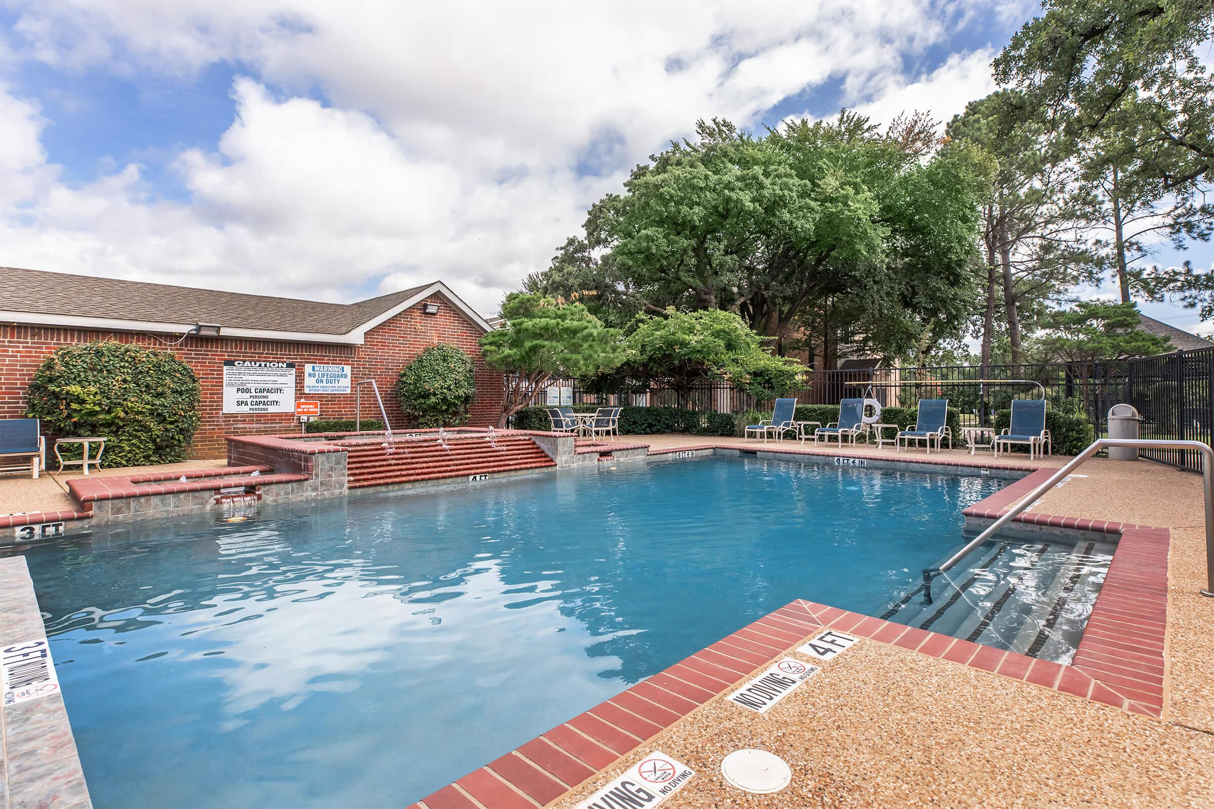 A clear swimming pool surrounded by a patio area, featuring lounge chairs and shaded trees. A brick wall is visible in the background, along with signs indicating pool rules and depth markers along the edges. The sky is partly cloudy, providing a bright atmosphere.