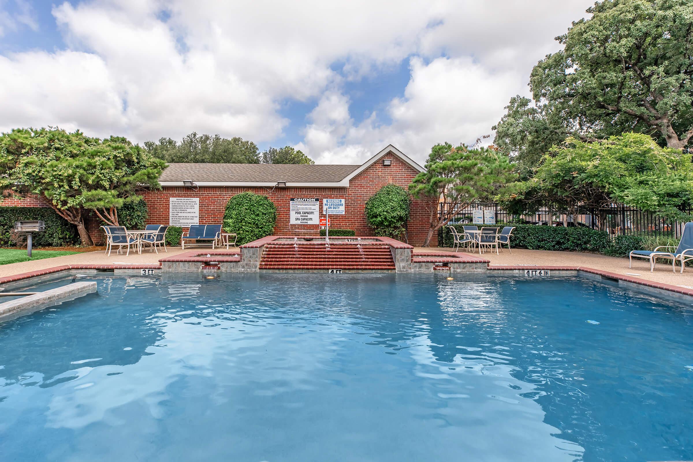 A clear blue swimming pool surrounded by lounge chairs and lush greenery, with a red brick building in the background. The sky is partly cloudy, adding to the sunny atmosphere. There are steps leading into the pool, creating an inviting space for relaxation and recreation.