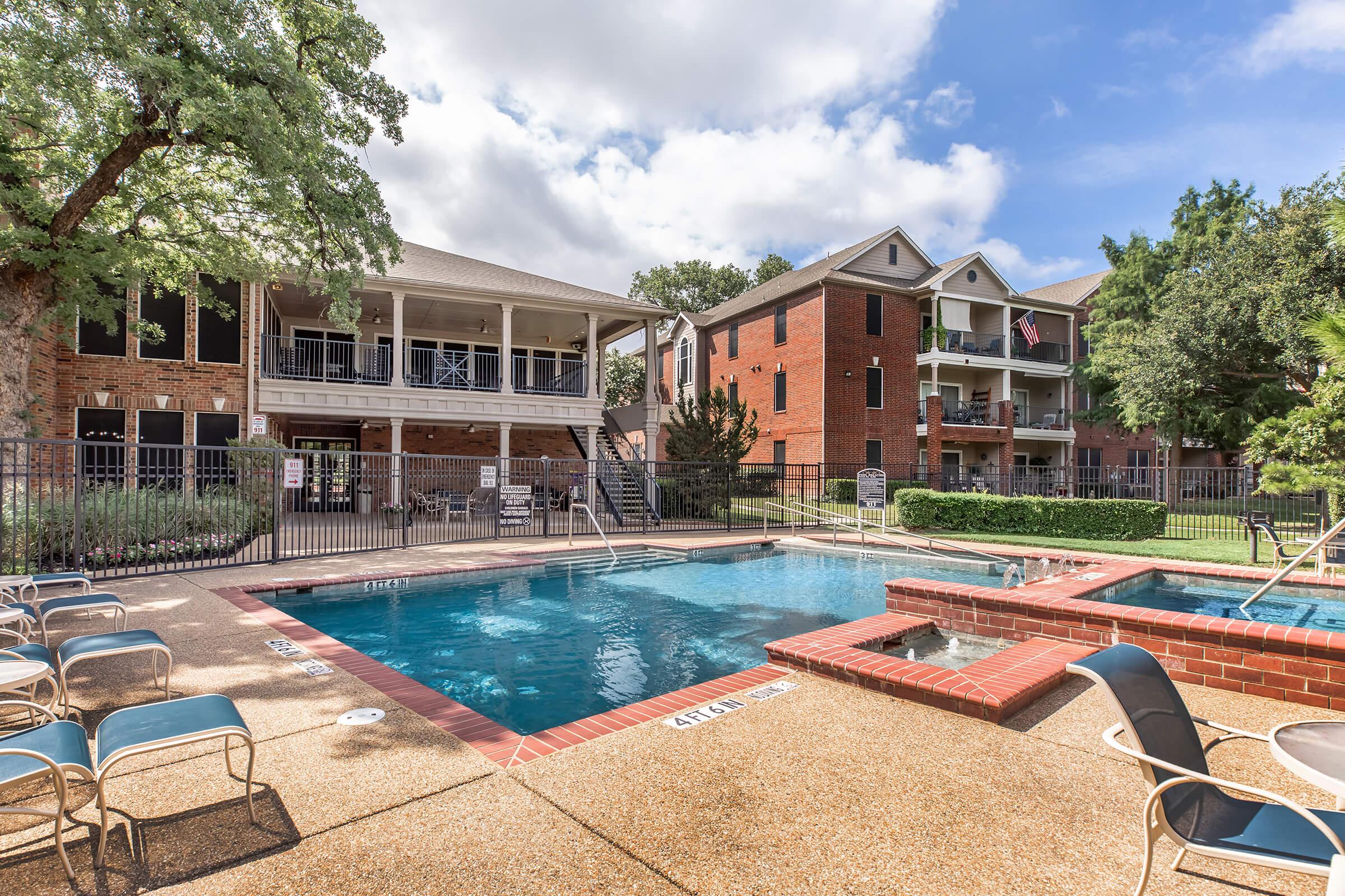 A well-maintained outdoor swimming pool with lounge chairs around it, surrounded by a fence. In the background, there are two residential buildings—one with a porch and the other with balconies. Lush greenery adds to the pleasant atmosphere, under a partly cloudy sky.